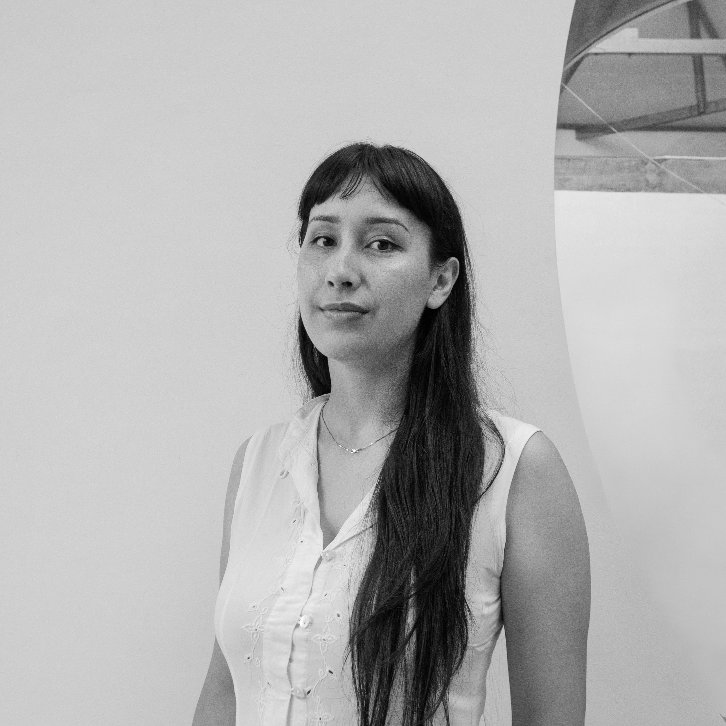 A woman with long dark hair and a sleeveless white blouse, standing against a plain wall with a modern architectural structure in the background.