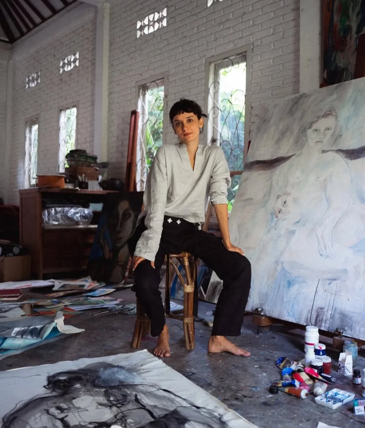 A woman sitting on a wooden stool in an art studio surrounded by paintings, art supplies, and sketches, with a large painting of a woman and a dog behind her.