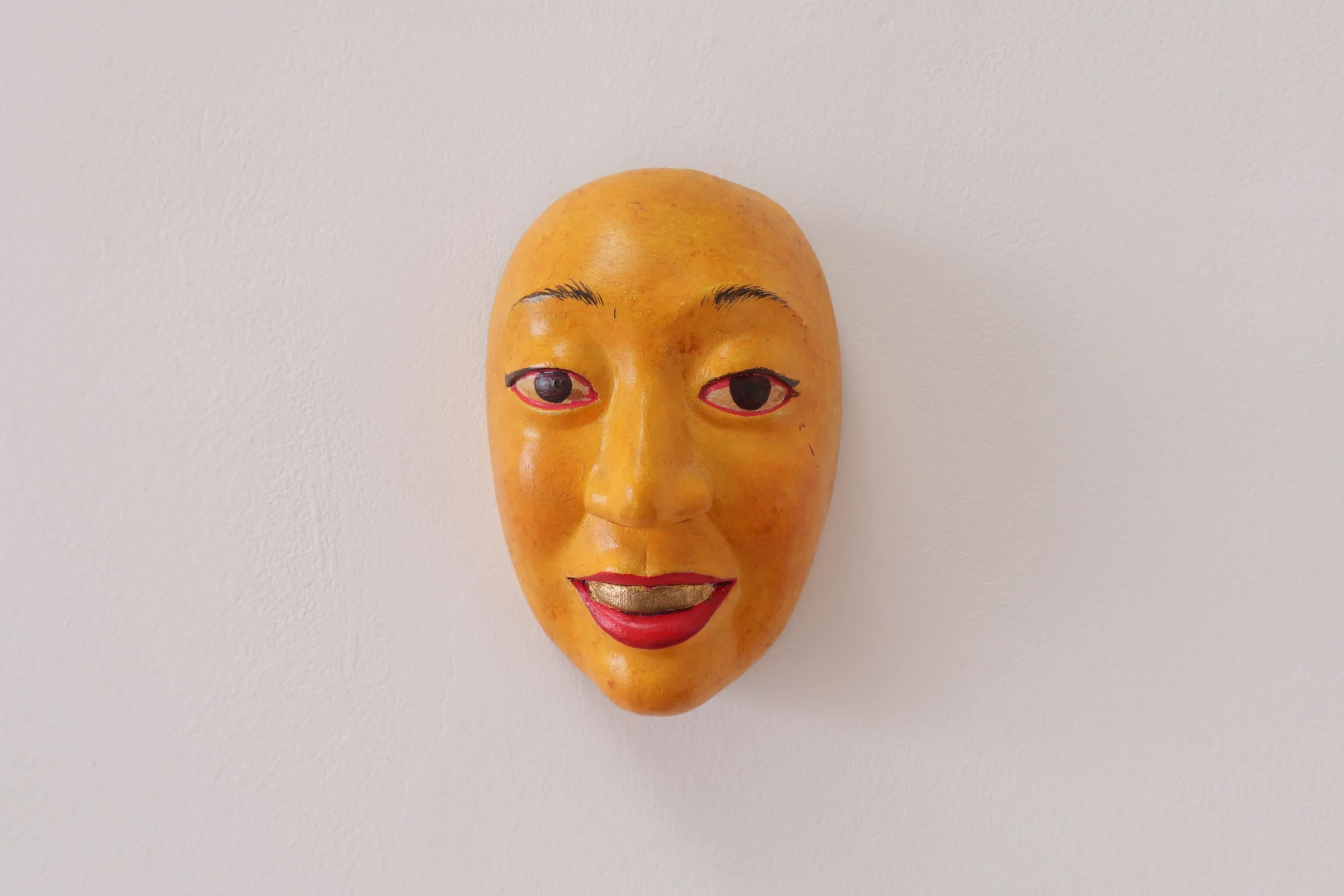 An orange-colored mask with human facial features, including painted eyes, eyebrows, nose, and mouth, mounted on a white wall.