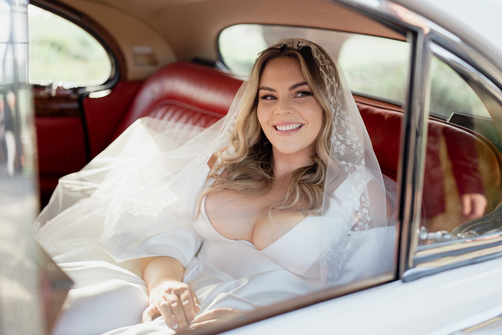 Bride sitting in a wedding dress in a vintage car, smiling.