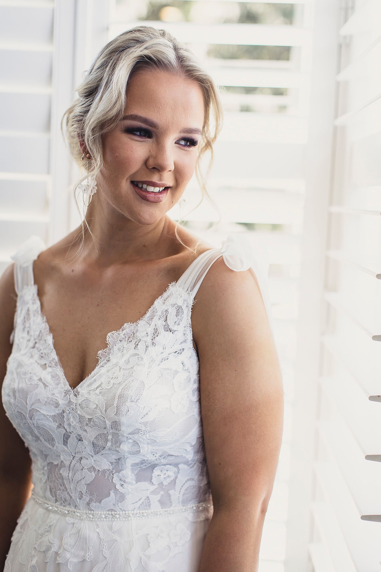 Bride in lace wedding dress looking out window with blinds.