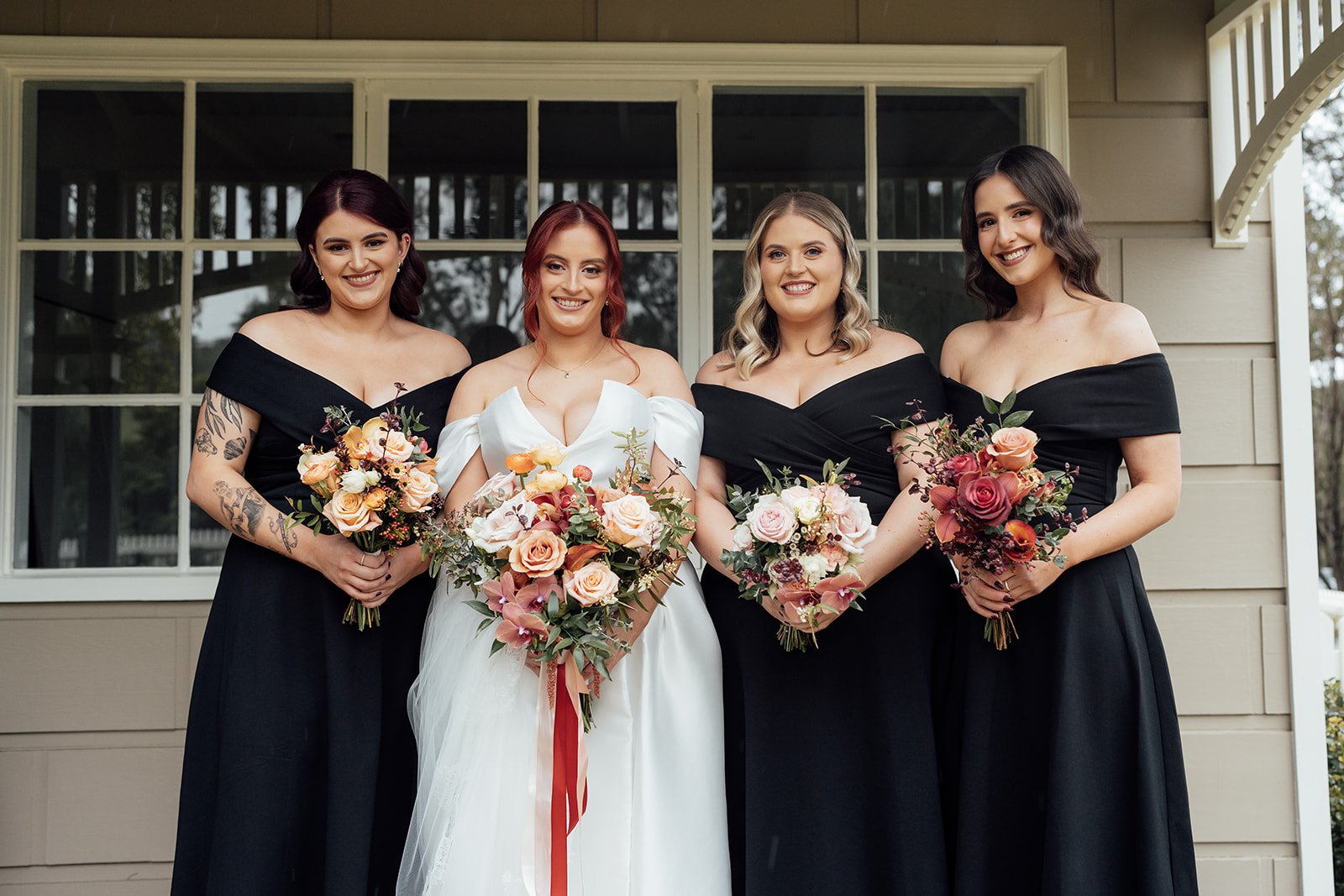 A bride in a white wedding dress stands with three bridesmaids wearing black off-the-shoulder dresses. They each hold floral bouquets with roses and greenery, posing in front of a window.