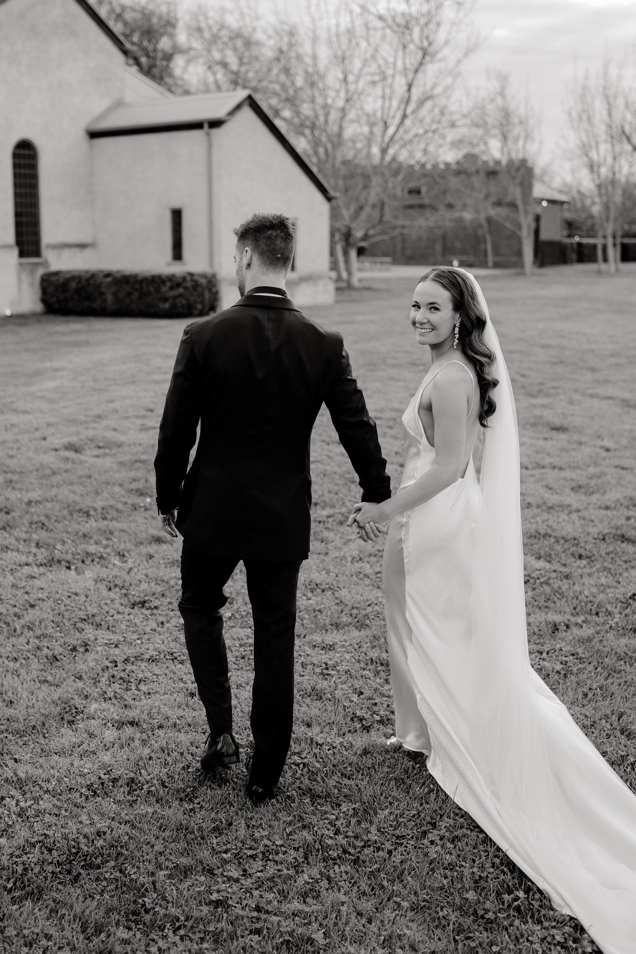 Bride and groom walking hand in hand on grass near a building, bride smiling back, wearing a long veil and gown, black and white photo.