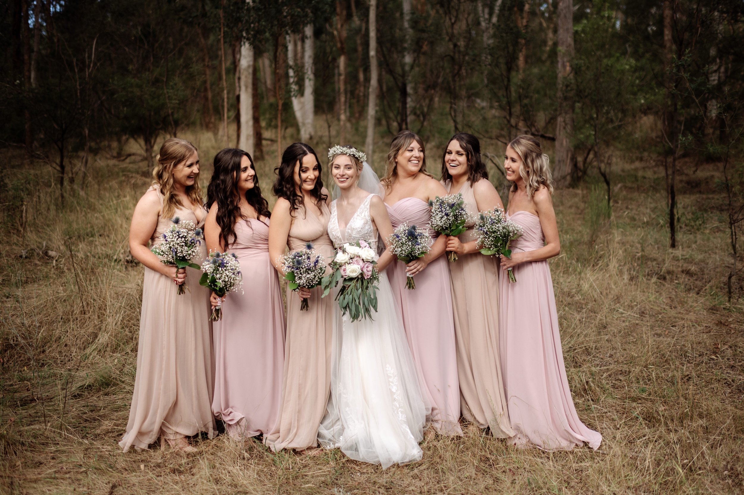 Bride with bridesmaids in pastel dresses holding bouquets in a forest setting.