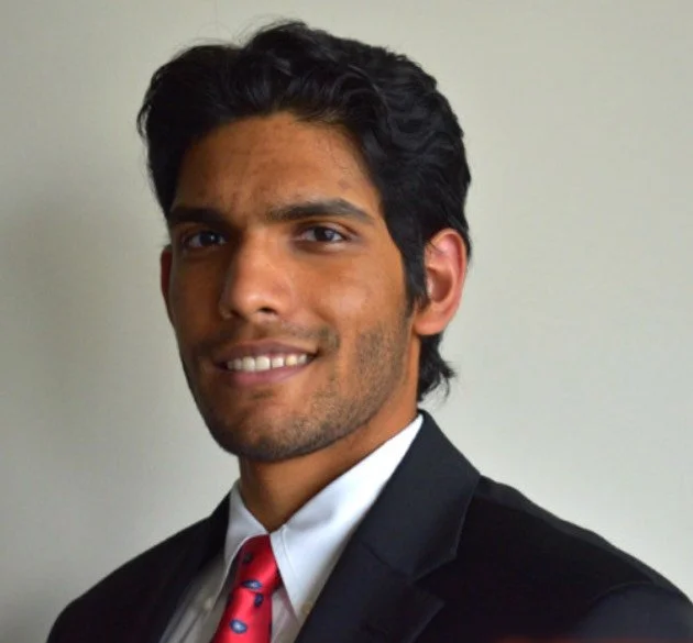 Headshot of a young man with dark hair, wearing a black suit, white shirt, and red tie, smiling at the camera against a plain background.