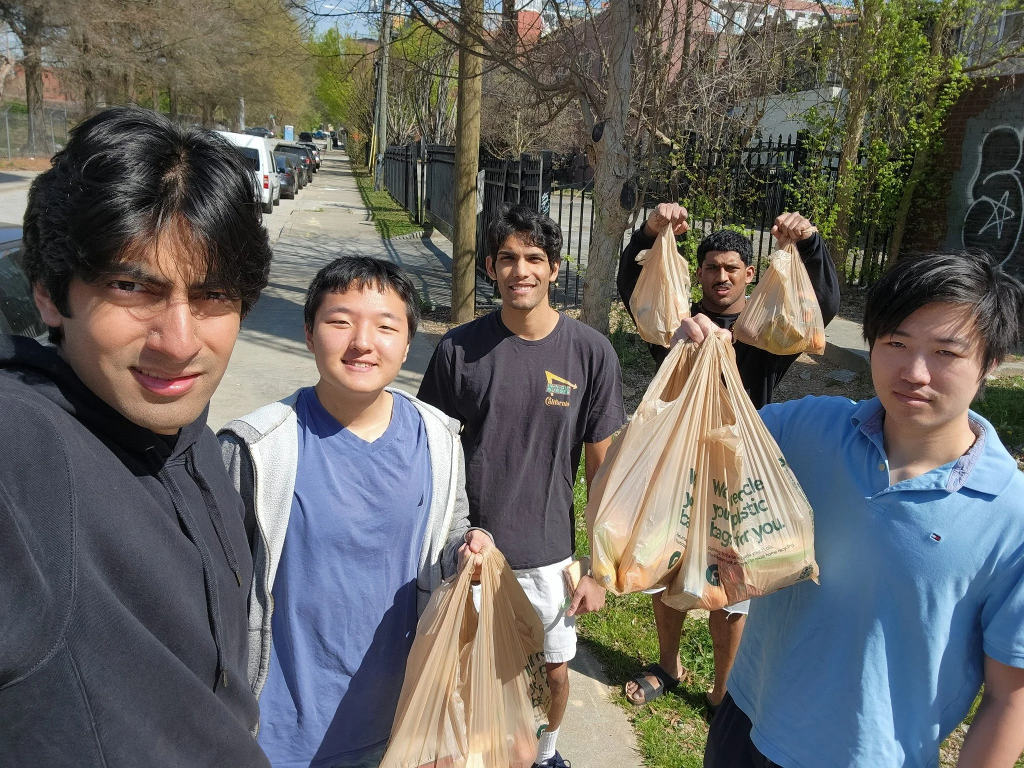 A group of five young men standing on a sidewalk, holding plastic bags filled with trash, likely participating in a community cleanup activity on a sunny day.