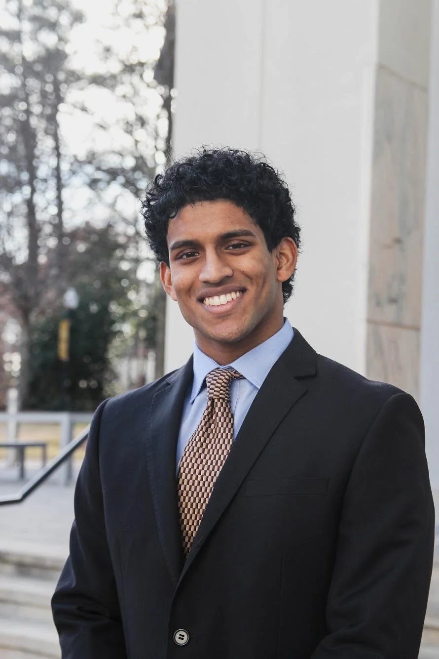A young man with curly black hair smiling outdoors, wearing a black suit, light blue shirt, and patterned tie.