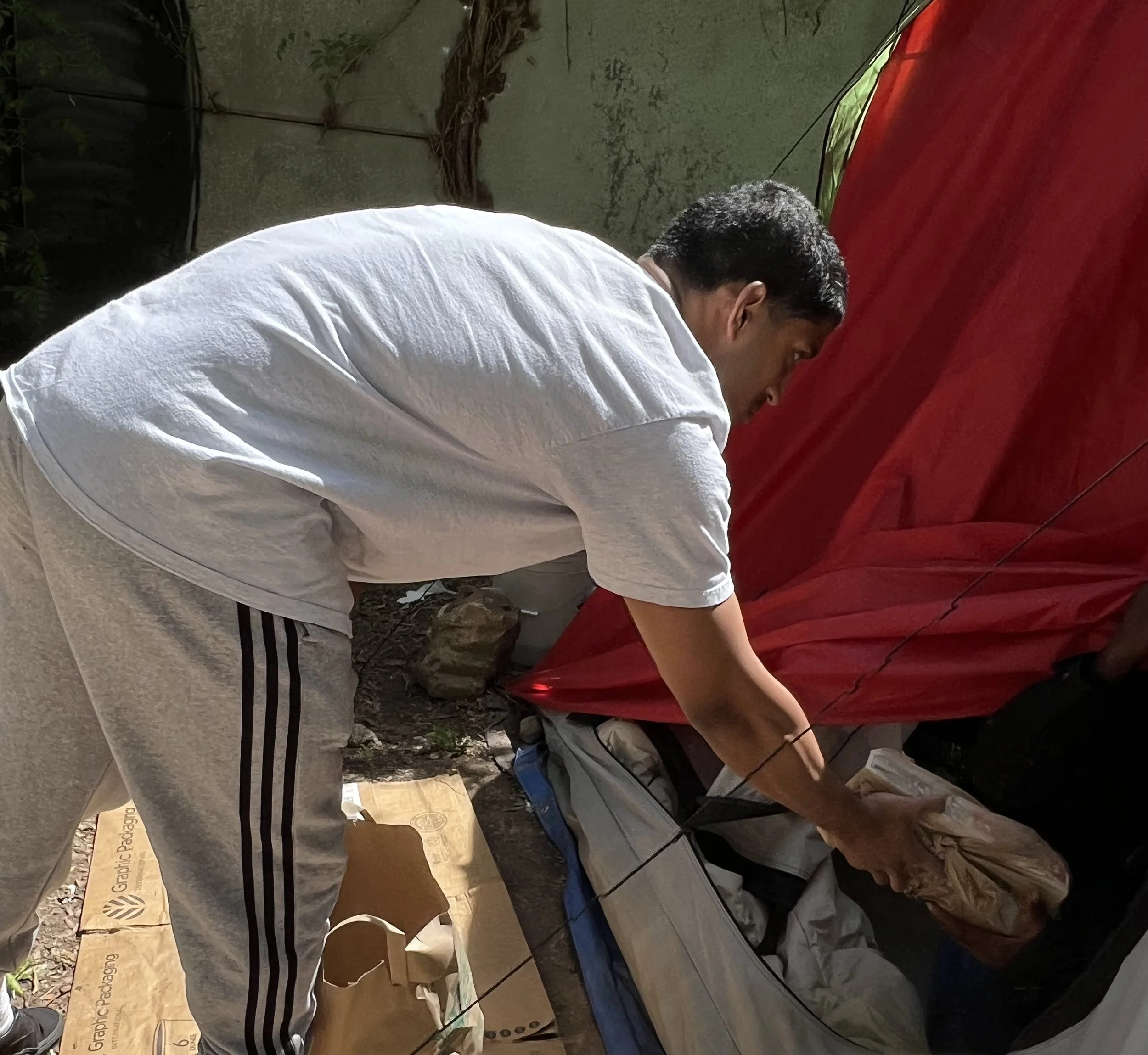 A man in a white t-shirt and grey sweatpants with black stripes is bending over, packing or unpacking items from a grey and blue tent that is set up outdoors. The tent has red fabric or cover, and there are cardboard boxes and bags around him. The setting appears to be a backyard or outdoor space with a concrete wall and some plants.