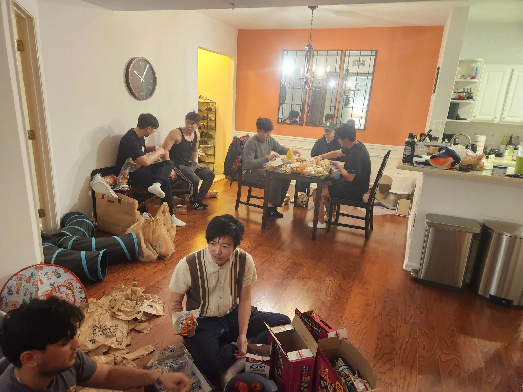 A group of young people gather in a living room and kitchen area, sitting around a table and on the floor. The scene appears casual and social, with some snacks and bags visible around them.
