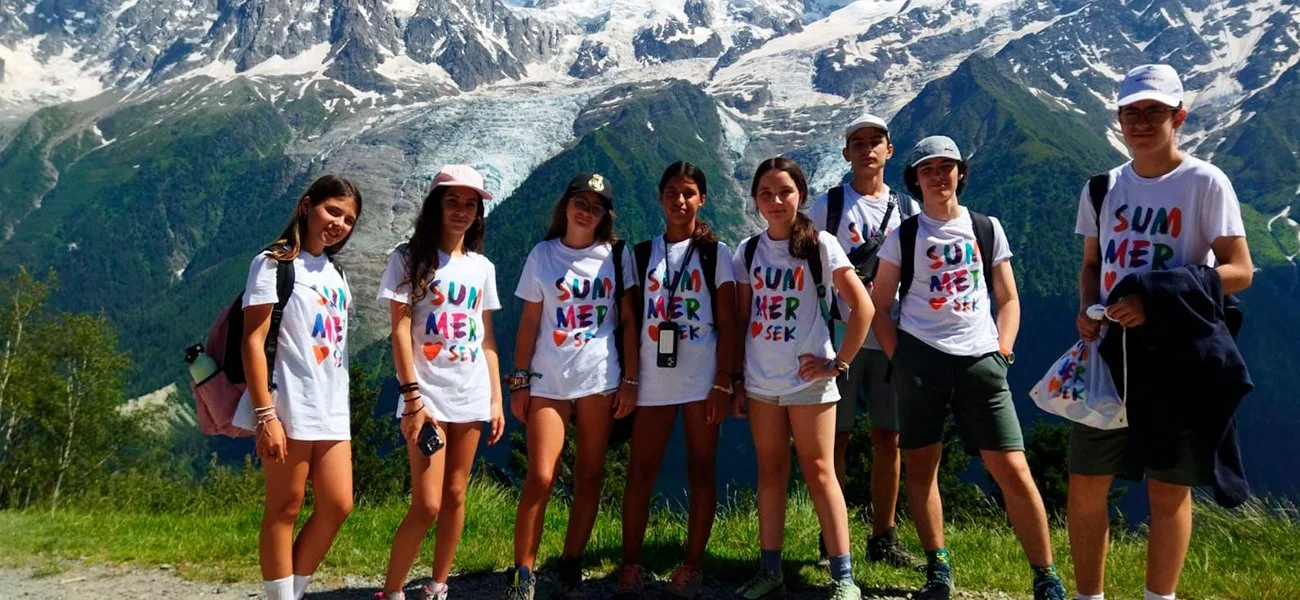 Group of eight young people standing outdoors in front of snow-capped mountains and green trees, wearing matching white T-shirts with colorful 'SUMMER' text, backpacks, and casual shorts or pants.