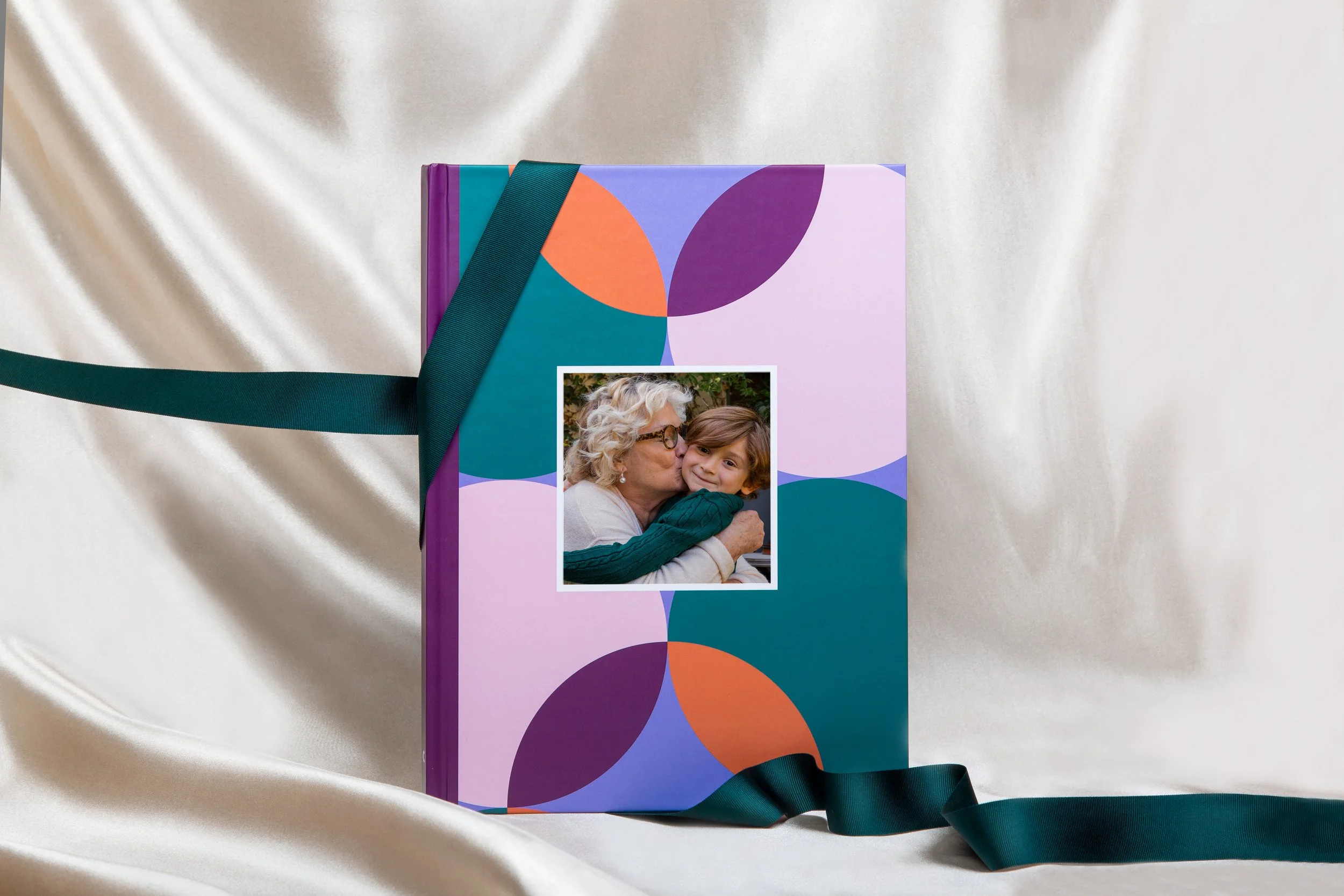 Livre avec un motif coloré, une photo d'une femme âgée embrassant un jeune garçon, posé sur un tissu de satin beige, avec une bande en satin vert foncé autour du livre.