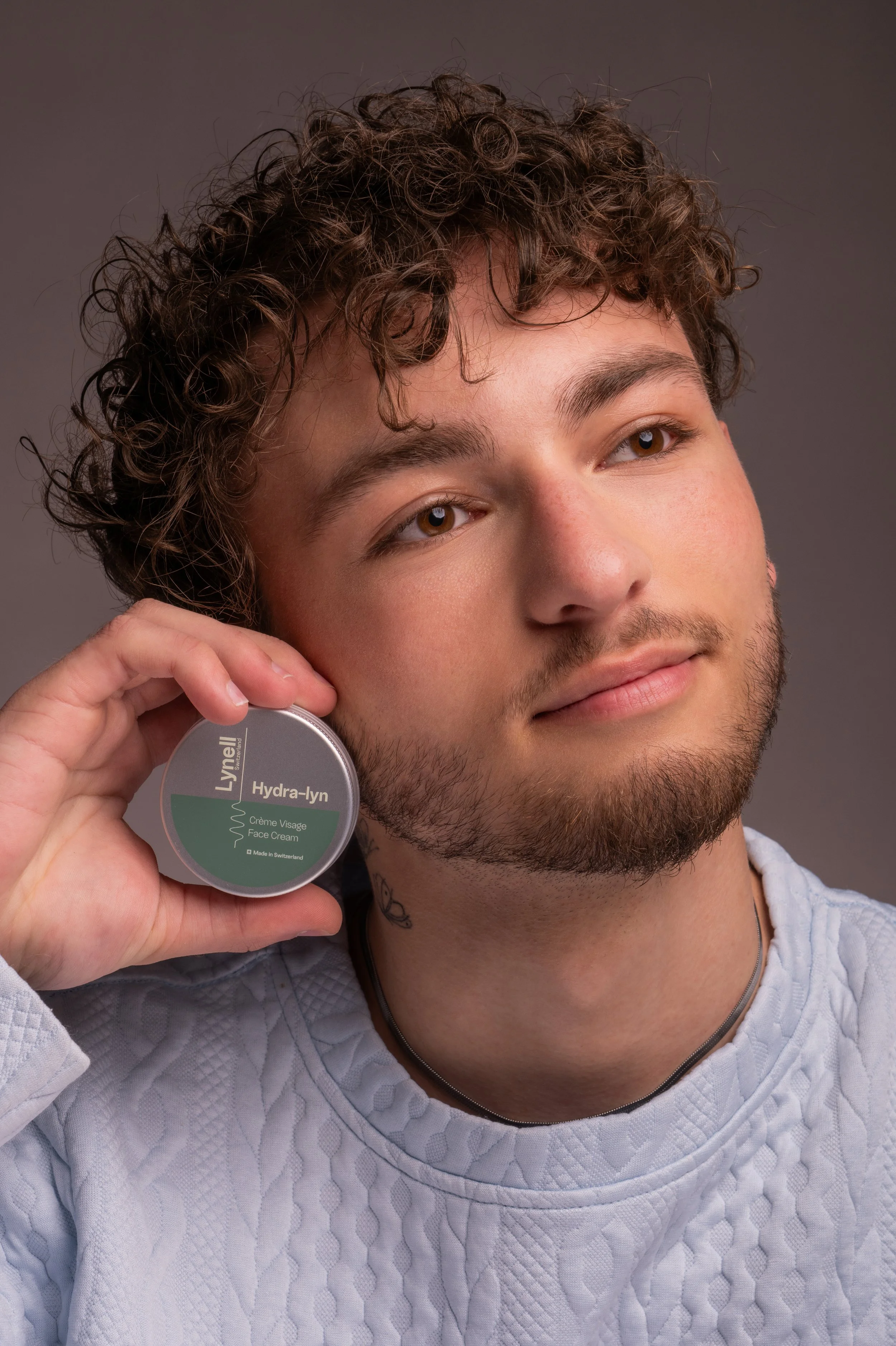 Un jeune homme avec des cheveux bouclés et une barbe, tenant un pot de crème de visage Lyvell Hydra-lyn