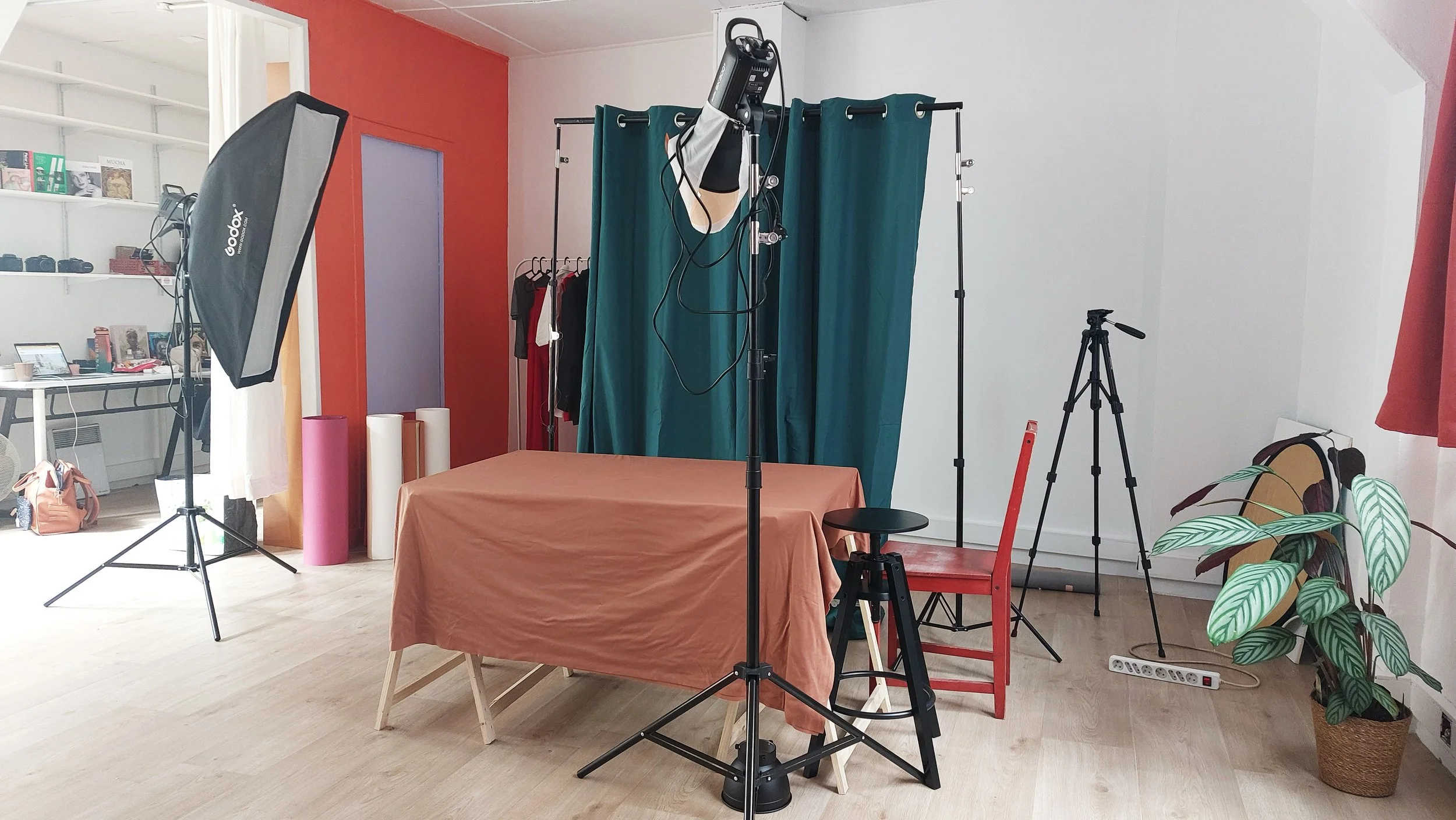 Studio photo avec table, chaises, éclairages, kit de photographie, plant, vêtements suspendus, et matériel de tournage.