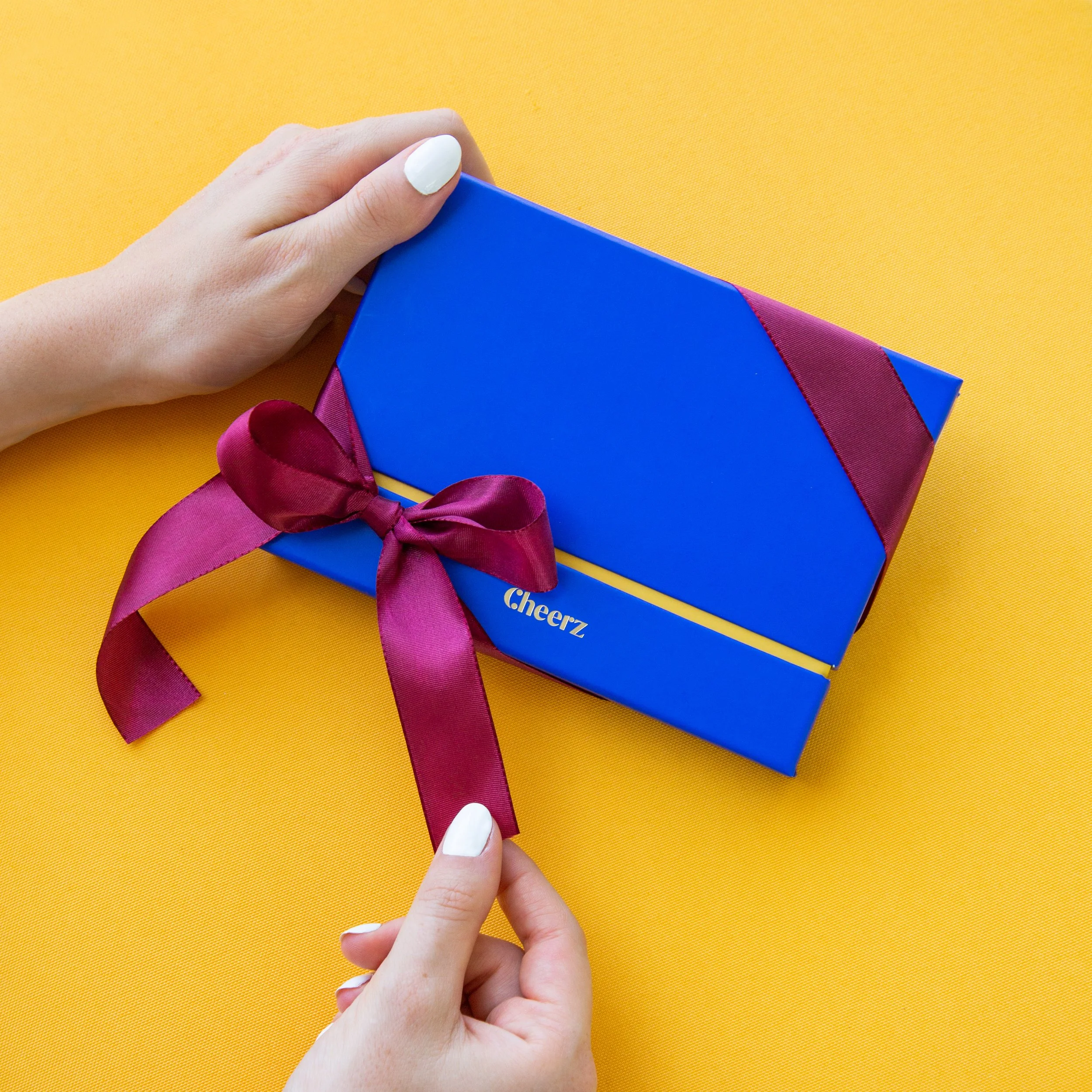 Une main tenant une boîte bleue avec un ruban rose, sur un fond jaune.