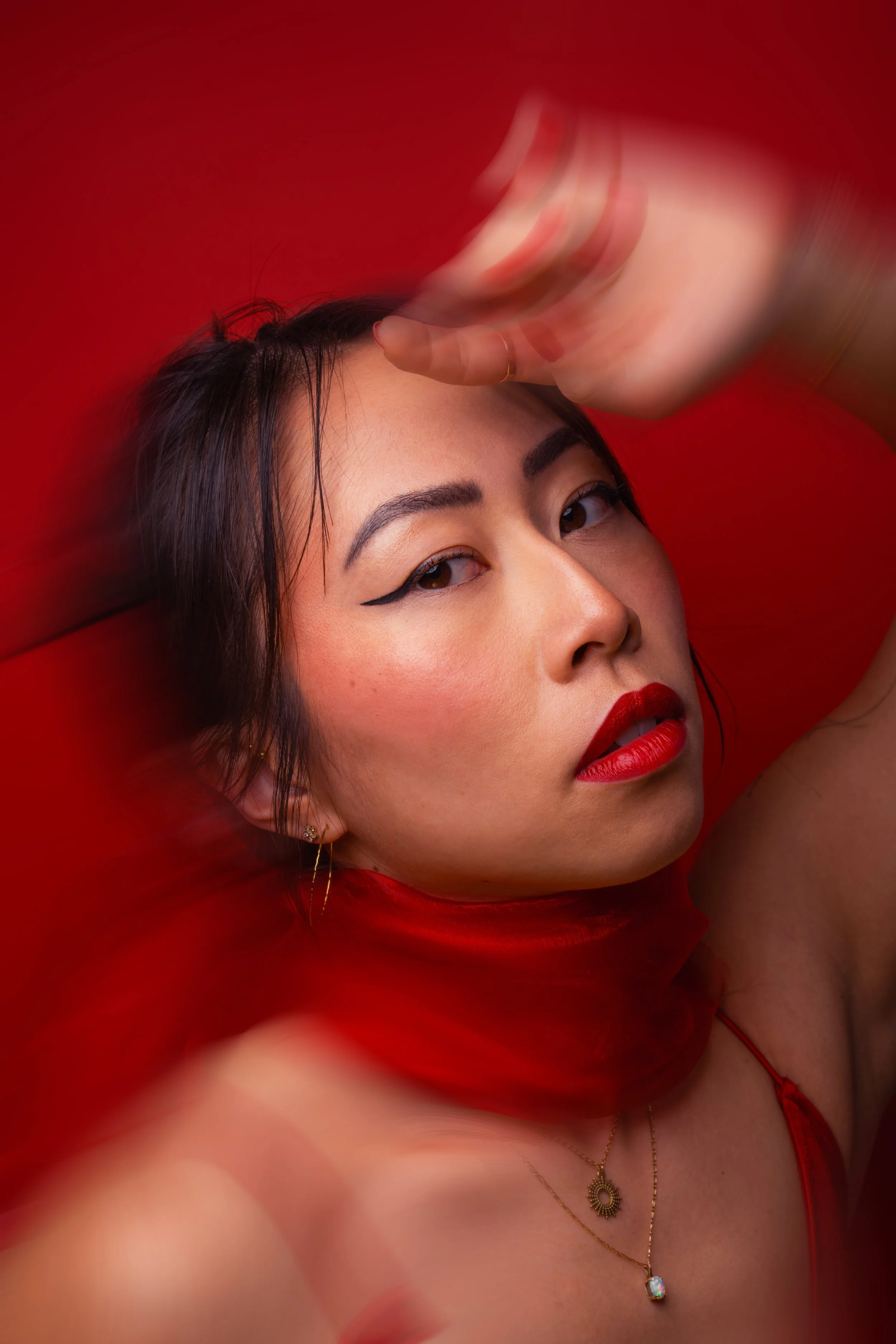 Jeune femme asiatique aux lèvres rouges, maquillage retroussé, portant une robe rouge et des bijoux or, posant sur un fond rouge.