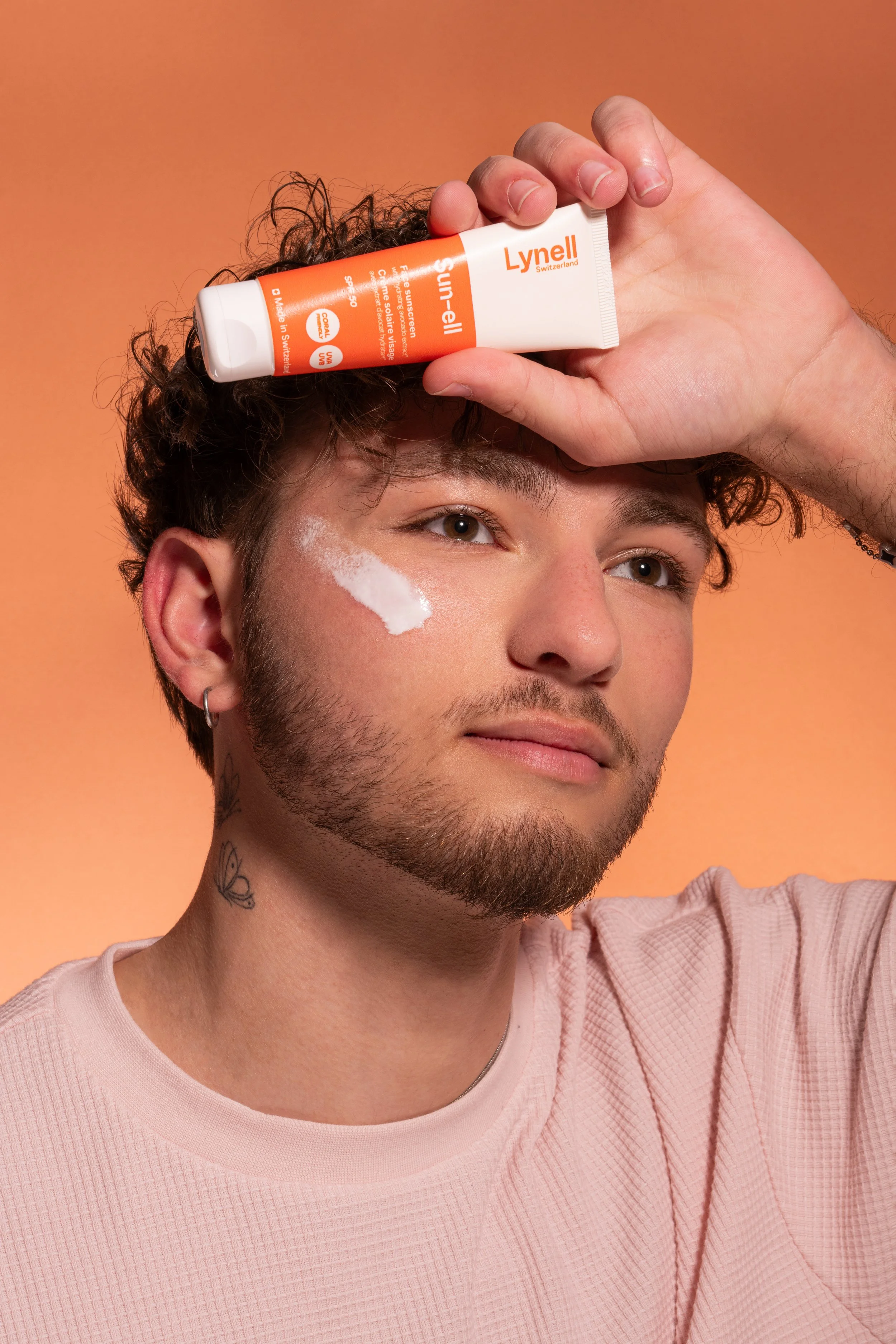 Un jeune homme aux cheveux bouclés et barbe courte, portant un T-shirt rose clair, tenant un tube de crème solaire Lynell au-dessus de son front, avec un peu de crème appliquée sur sa joue, contre un fond orange.