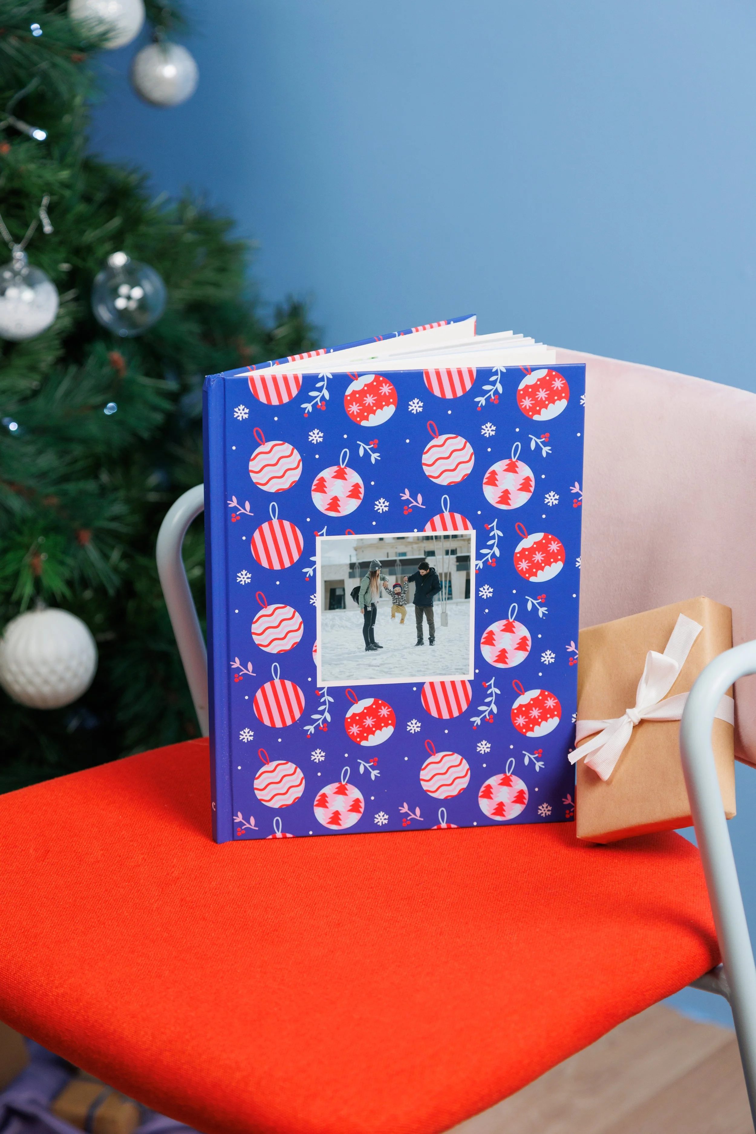 Un livre de Noël avec une couverture bleue décorée de motifs de boules de Noël rouges et blanches. Sur la couverture, une photo de famille sur la glace enneigée. Un cadeau enveloppé de papier brun avec un ruban blanc repose à côté, sur une chaise ave