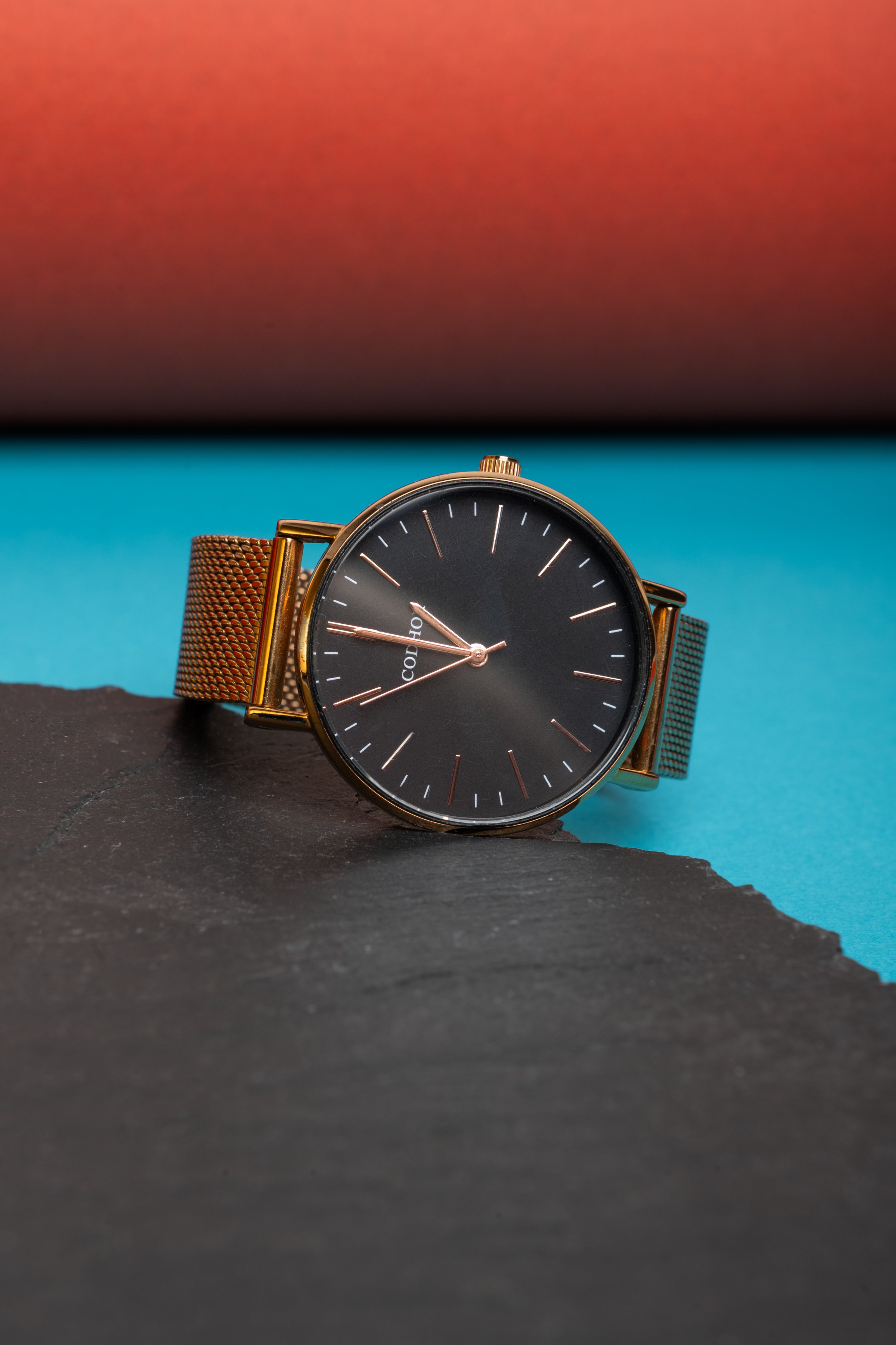 Montre à cadran noir avec bracelet en maille dorée posé sur une roche noire, fond bleu et dégradé rose en haut