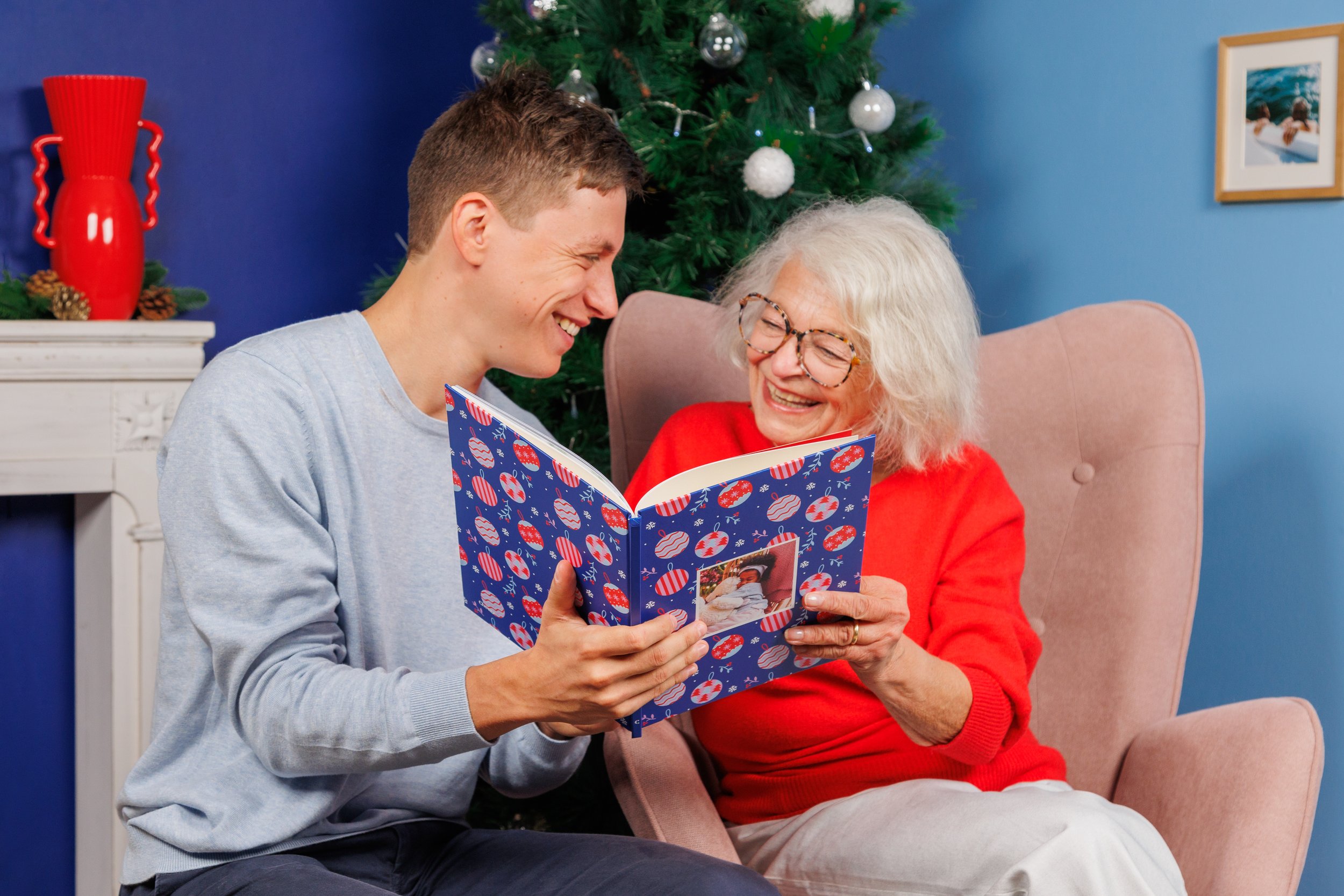 Un jeune homme et une femme âgée sourient en regardant un album de photos de Noël, dans une ambiance festive avec un sapin de Noël décoré en arrière-plan.
