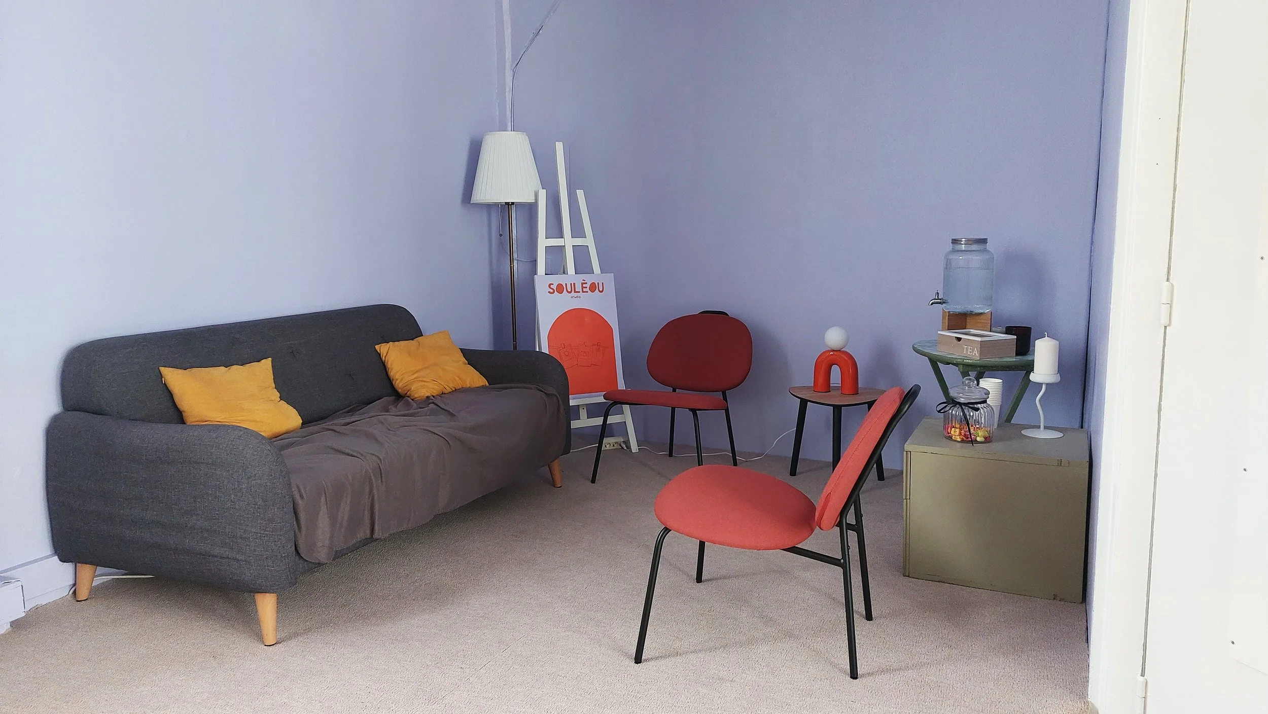 Salon avec un canapé gris, deux chaises rouges, une table noire, une lampe blanche, un poster orange, un petit meuble beige, un distributeur d'eau, un livre, une boîte, un récipient transparent avec des bonbons, et une lampe