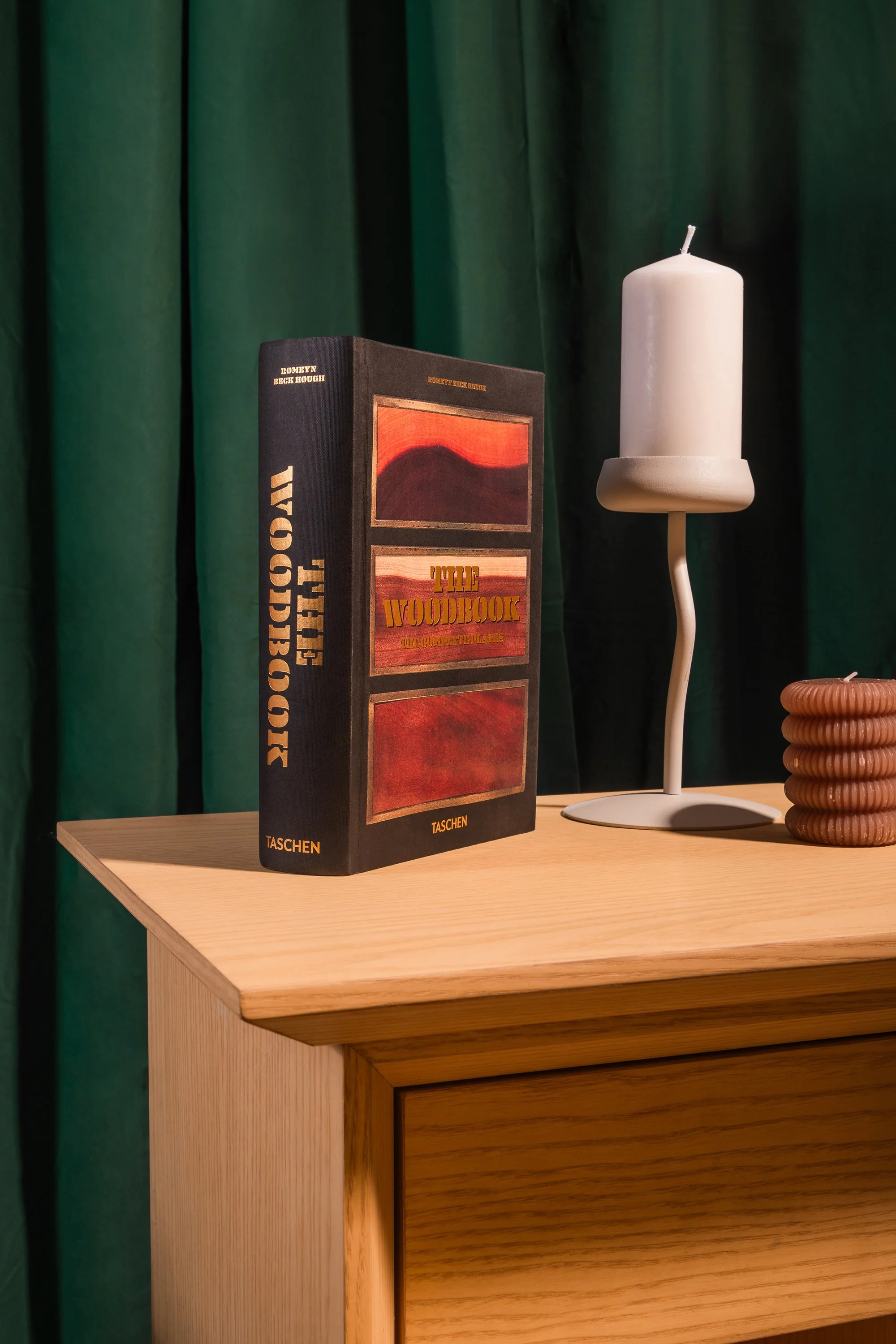 Un livre intitulé 'The Woodbook' posé sur une table en bois clair, à côté d'une bougie blanche sur un support incurvé et d'un objet décoratif en forme de rubans de couleur beige, avec un rideau vert en arrière-plan.