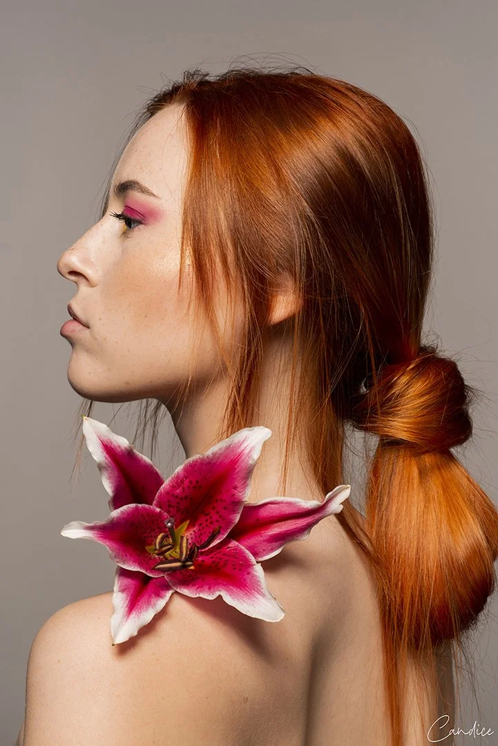 Une femme aux cheveux roux avec un grand lys rose sur son épaule, regardant vers la gauche, contre un fond gris