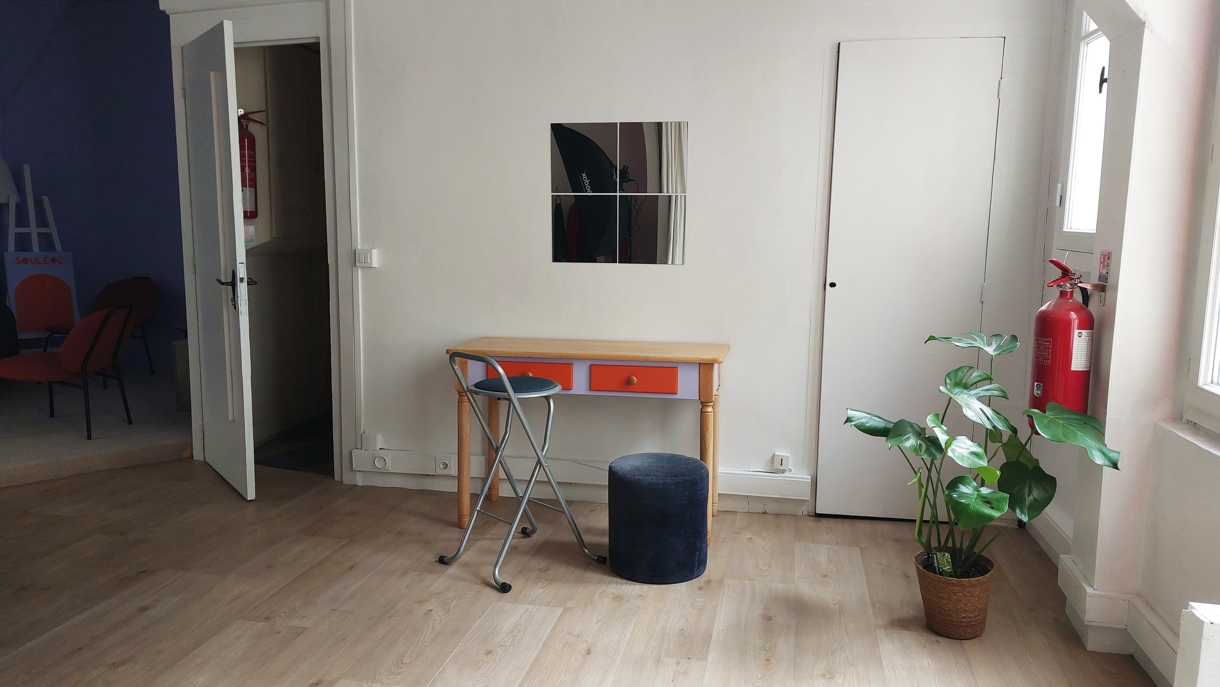Intérieur d'une pièce avec un petit bureau, un miroir, une lampe de studio, un tabouret, une plante verte en pot et un extincteur à proximité d'une fenêtre.
