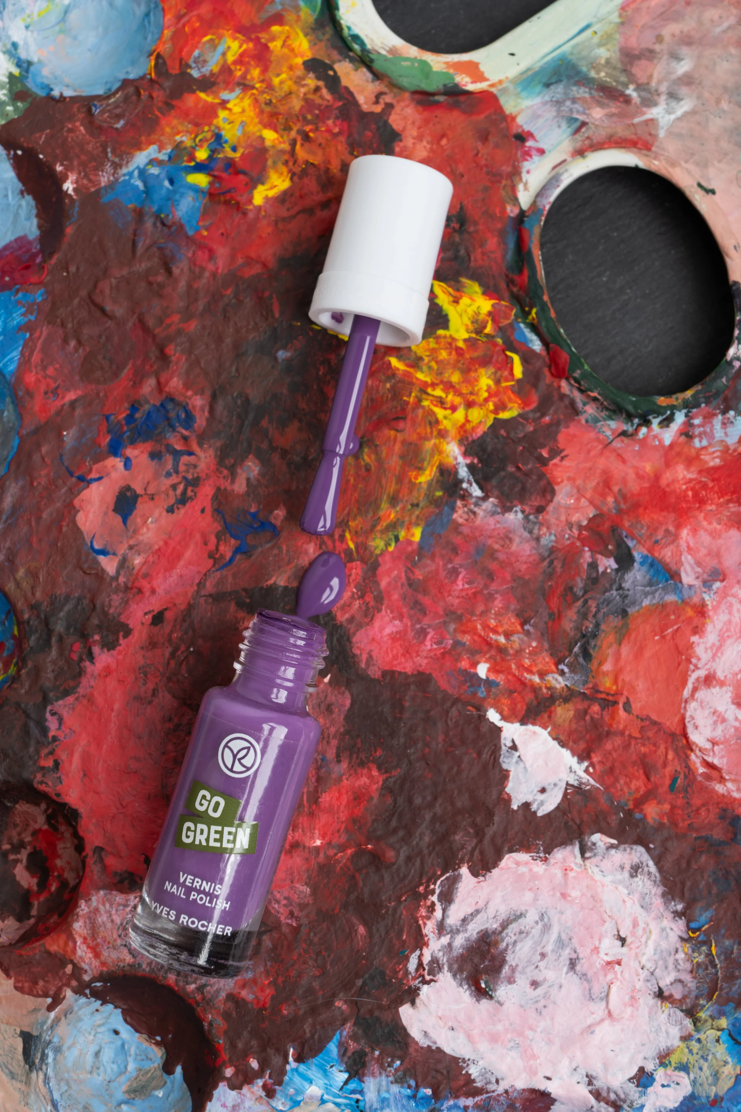 Vernis à ongles violet ouvert avec le pinceau posé sur une palette de peinture colorée.