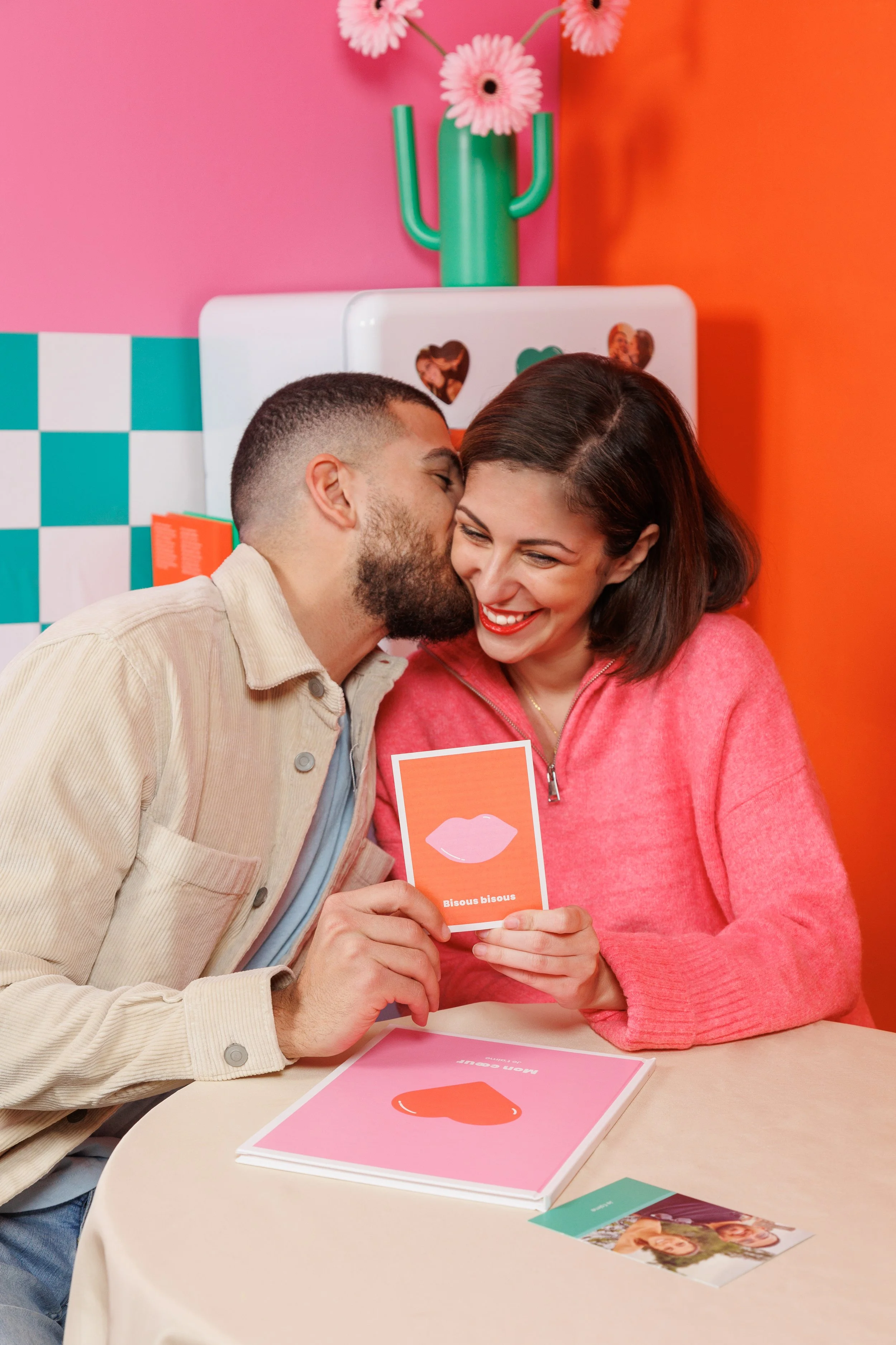 Un homme et une femme souriants partagent un moment affectueux, la femme tient une carte avec une illustration de lèvres et le texte 'Bisous bisous'.