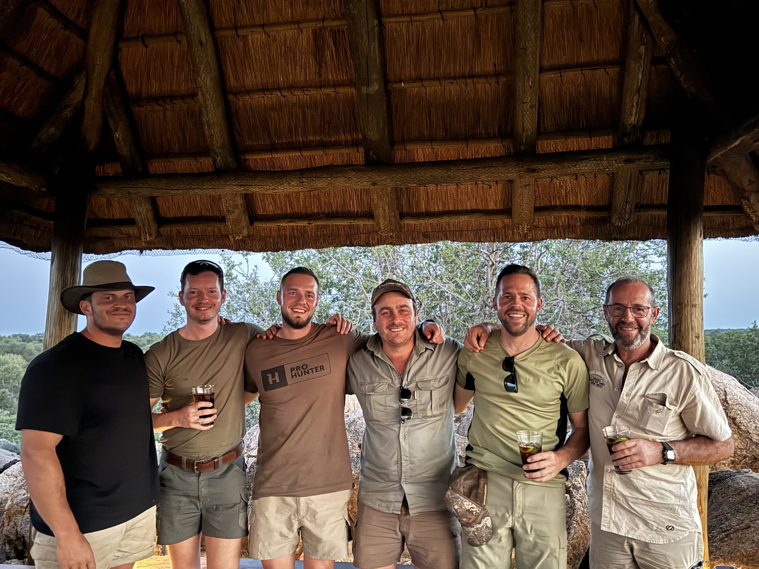 Group of seven men standing together under a thatched roof, smiling, with trees and rocks in the background, some holding drinks.