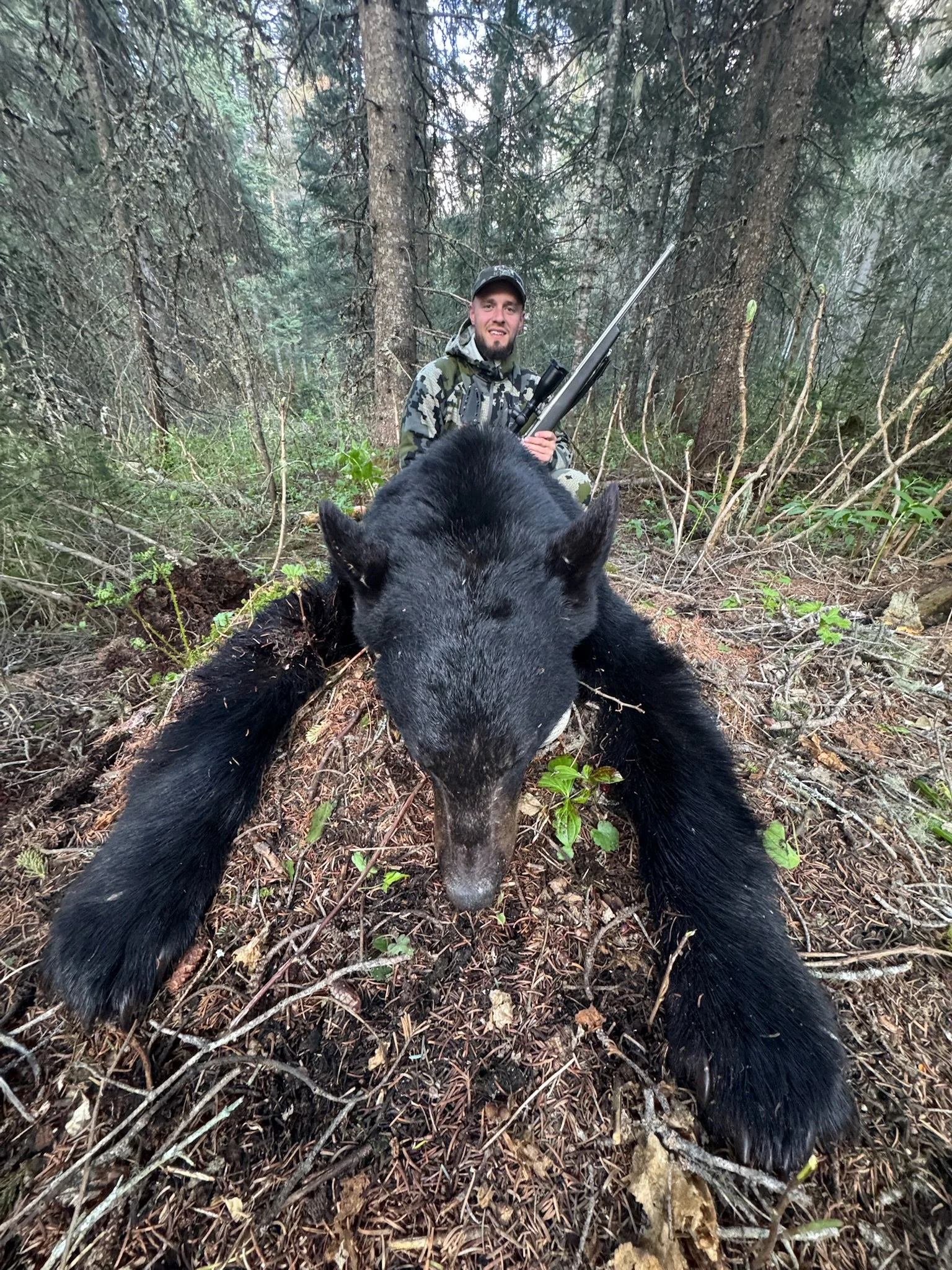 A man in camouflage clothing kneels behind a large black bear he has hunted, holding a rifle in a forested area with trees and underbrush.