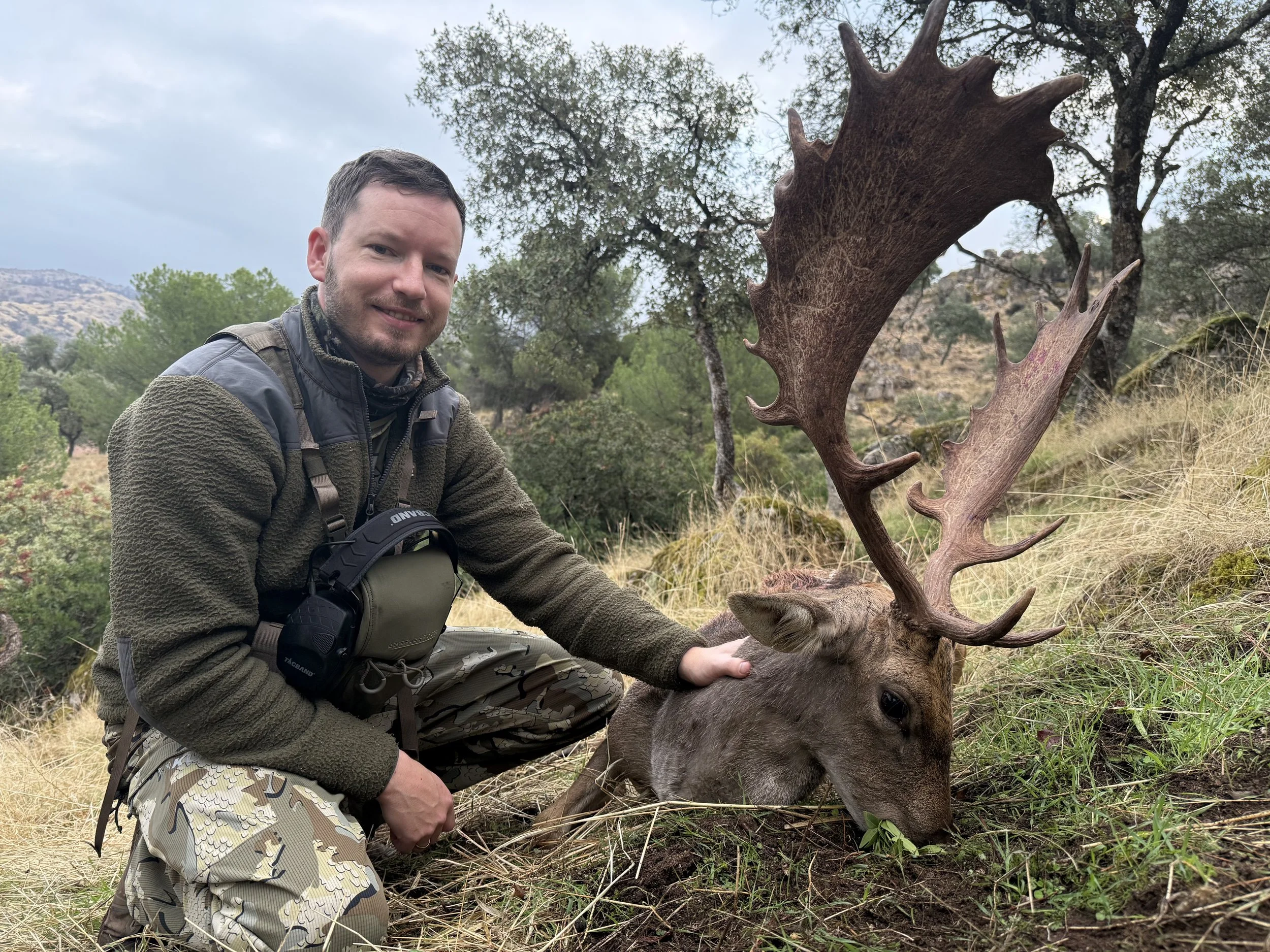 A man in camo pants and a jacket kneels next to a harvested deer with large antlers in a grassy outdoor setting