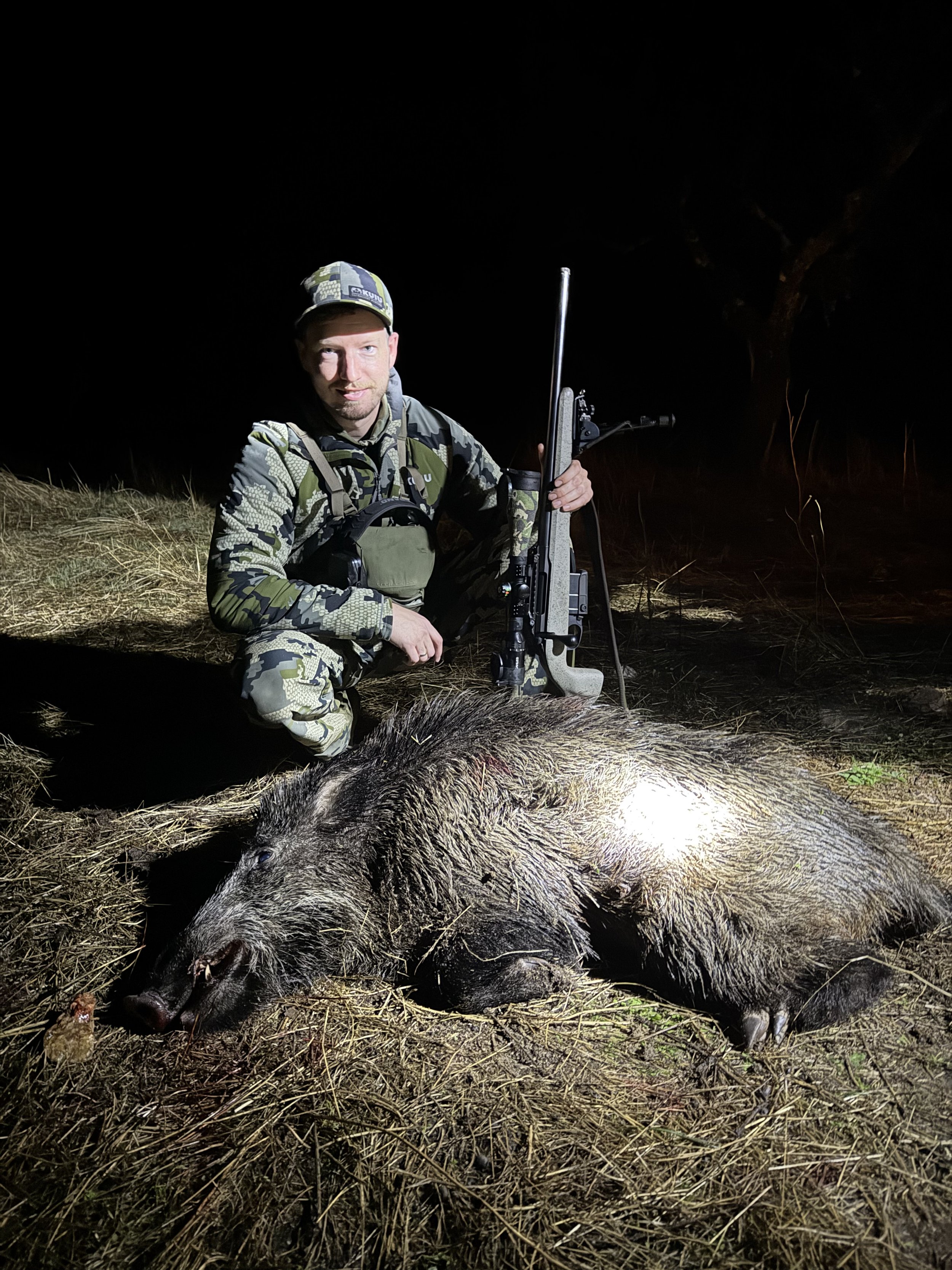Man in camouflage clothing kneeling next to a dead wild boar with a rifle at night.