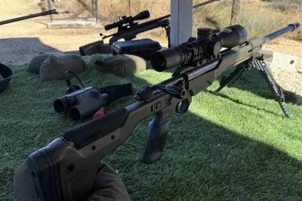 A modern bolt-action rifle with a scope and bipod, held by a person wearing gloves, set on a green artificial grassy surface outdoors.