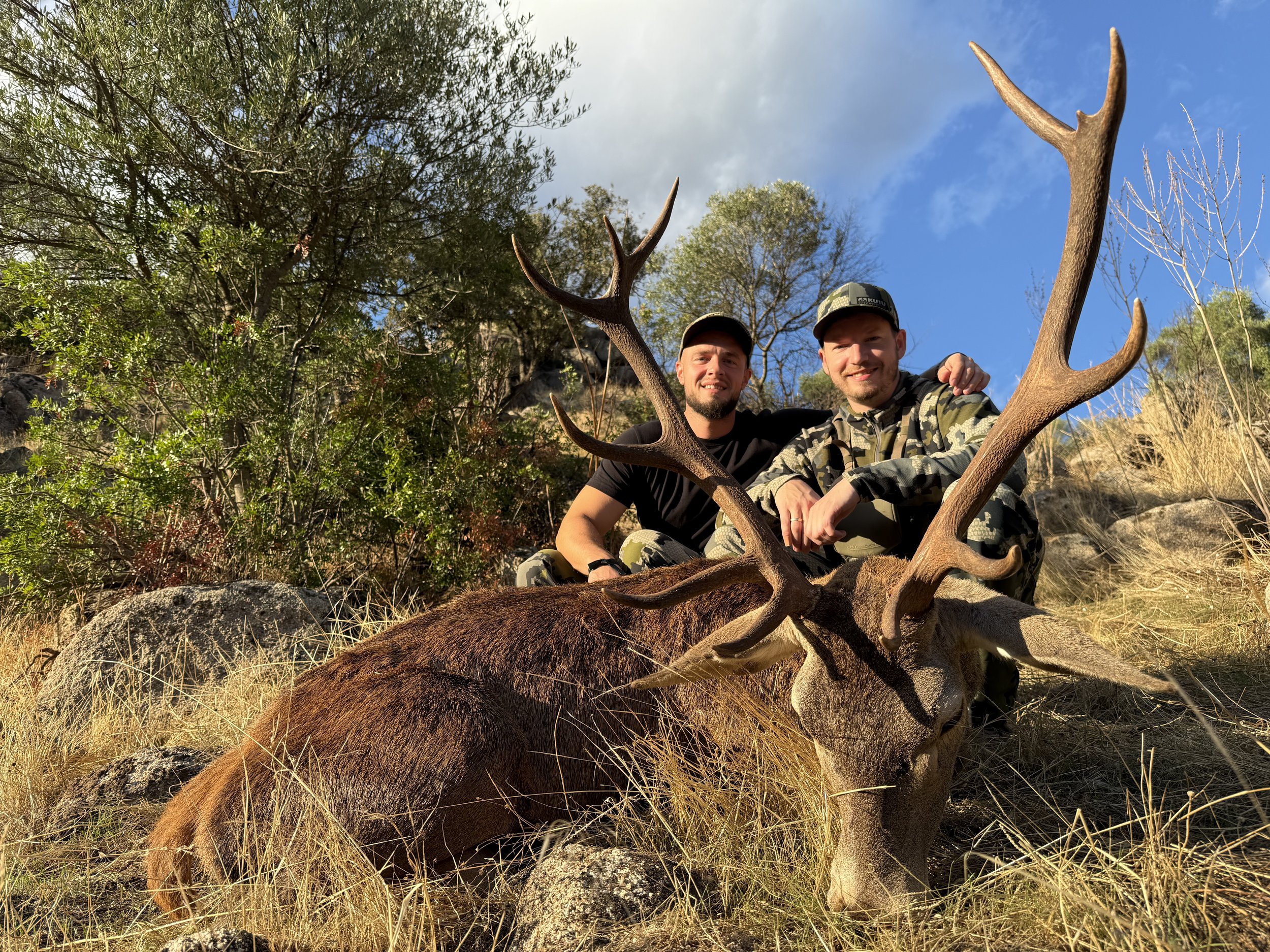 Two men in camouflage clothing and caps sit behind a large fallen deer with impressive antlers in a natural outdoor setting with rocks, dry grass, and trees.