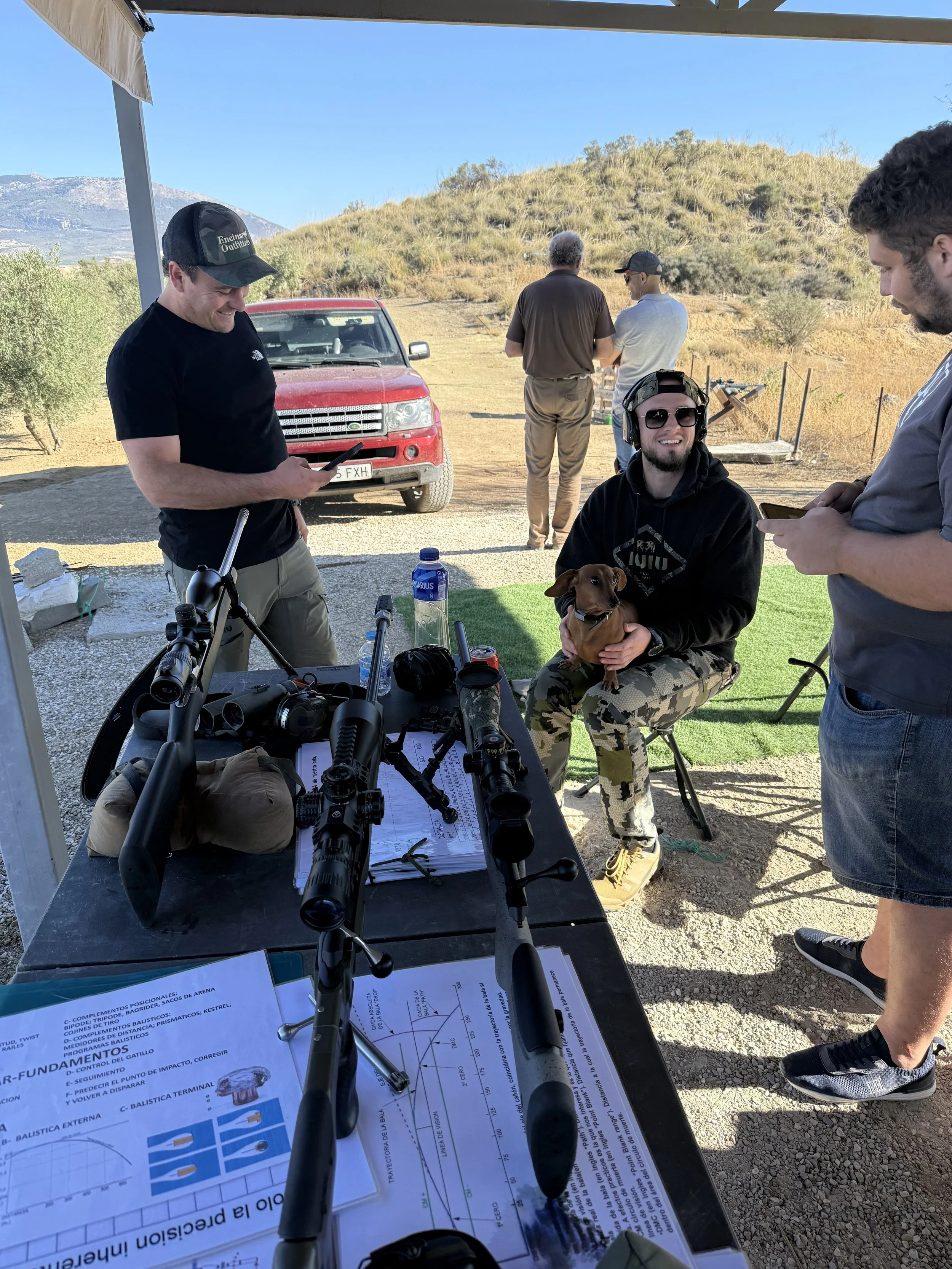 Group of people at outdoor shooting range with rifles and scopes, one person sitting with a small dog, others using their phones, set against a desert landscape with mountains in the background.