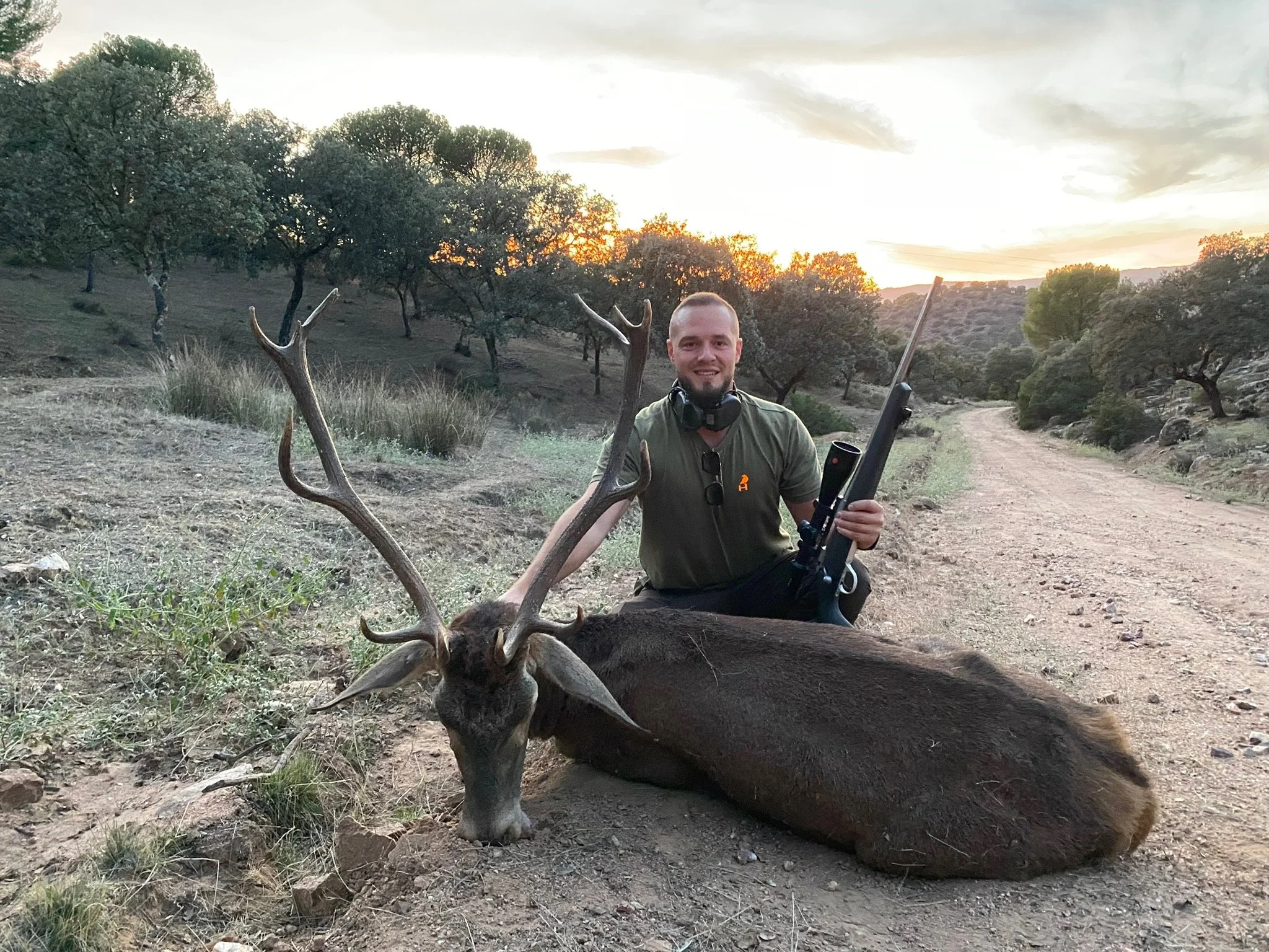 A man with a rifle kneels next to a large, freshly hunted deer with antlers, lying on the dirt road during sunset in a rural landscape with scattered trees.