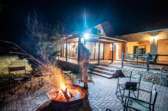 Person standing near a lit fire pit outside a house at night, with the house's large windows and a patio visible.