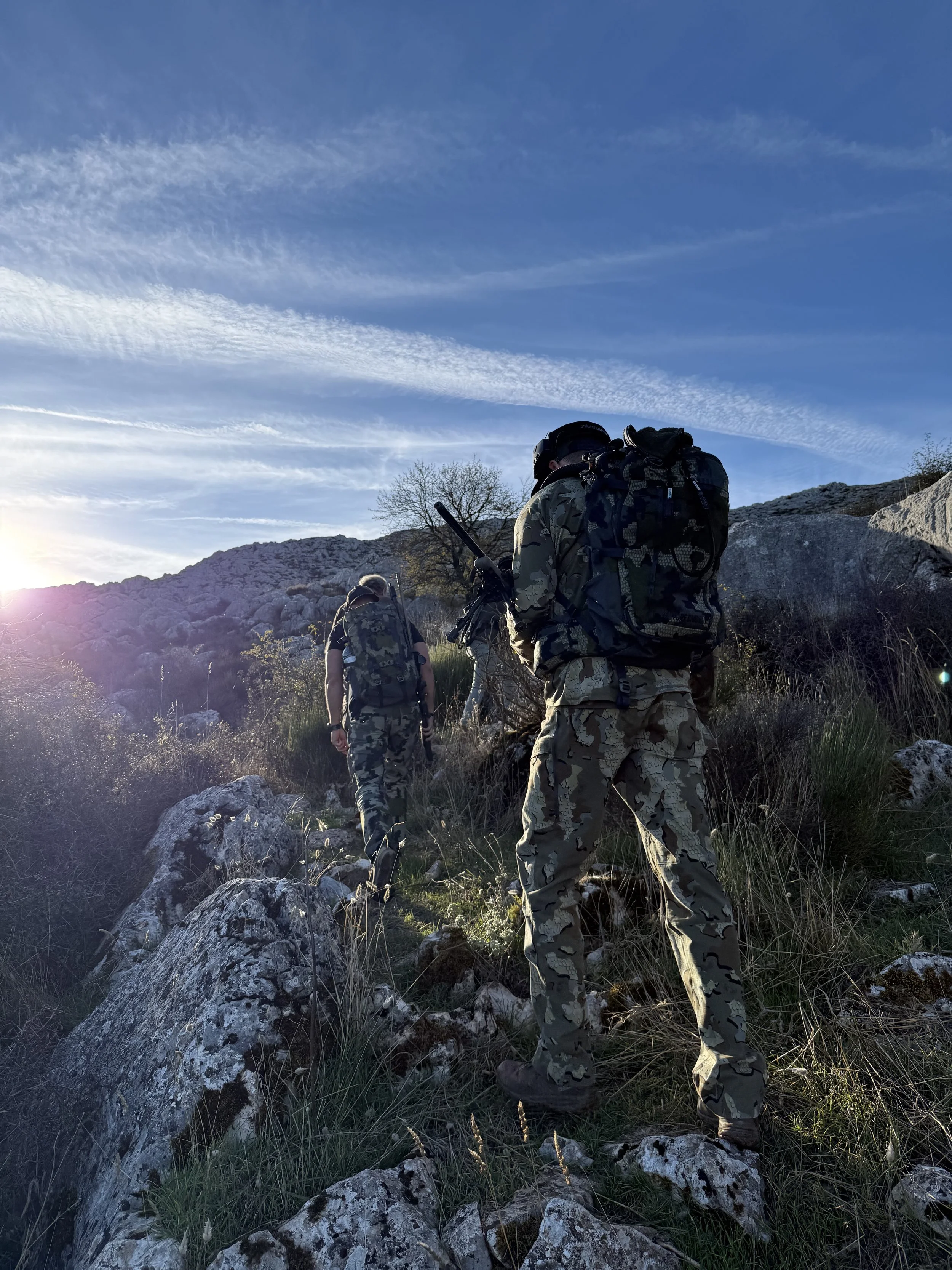 Three soldiers hiking up a rocky hill during sunset with backpacks, wearing camouflage uniforms and carrying gear.