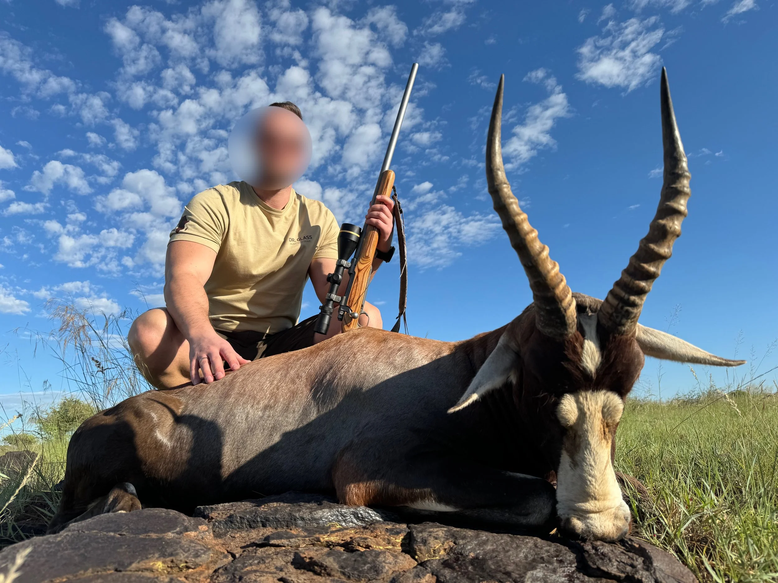 A person kneeling on grass in a field next to a lying antelope with large, spiral horns, under a partly cloudy blue sky.