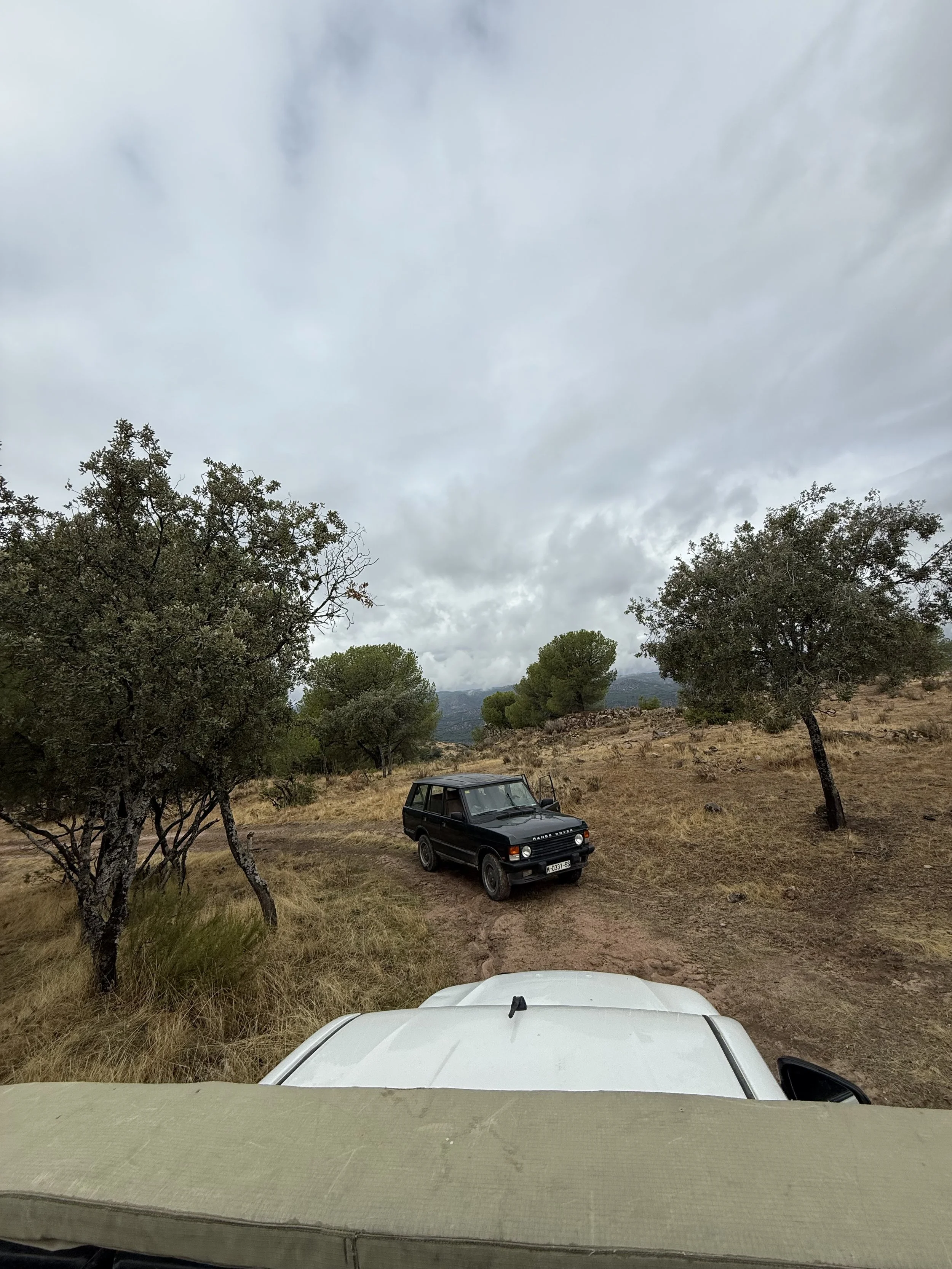 View from inside a vehicle looking out at a rough dirt trail with sparse grass and trees. A black SUV is parked on the trail, and more trees and hilly terrain are visible in the background under a cloudy sky.