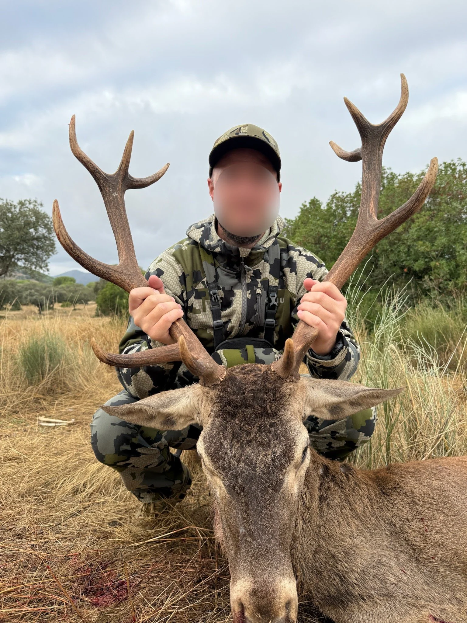 Person in camouflage hunting gear kneeling behind a large dead deer with antlers, holding the antlers in a field with grass and trees in the background.