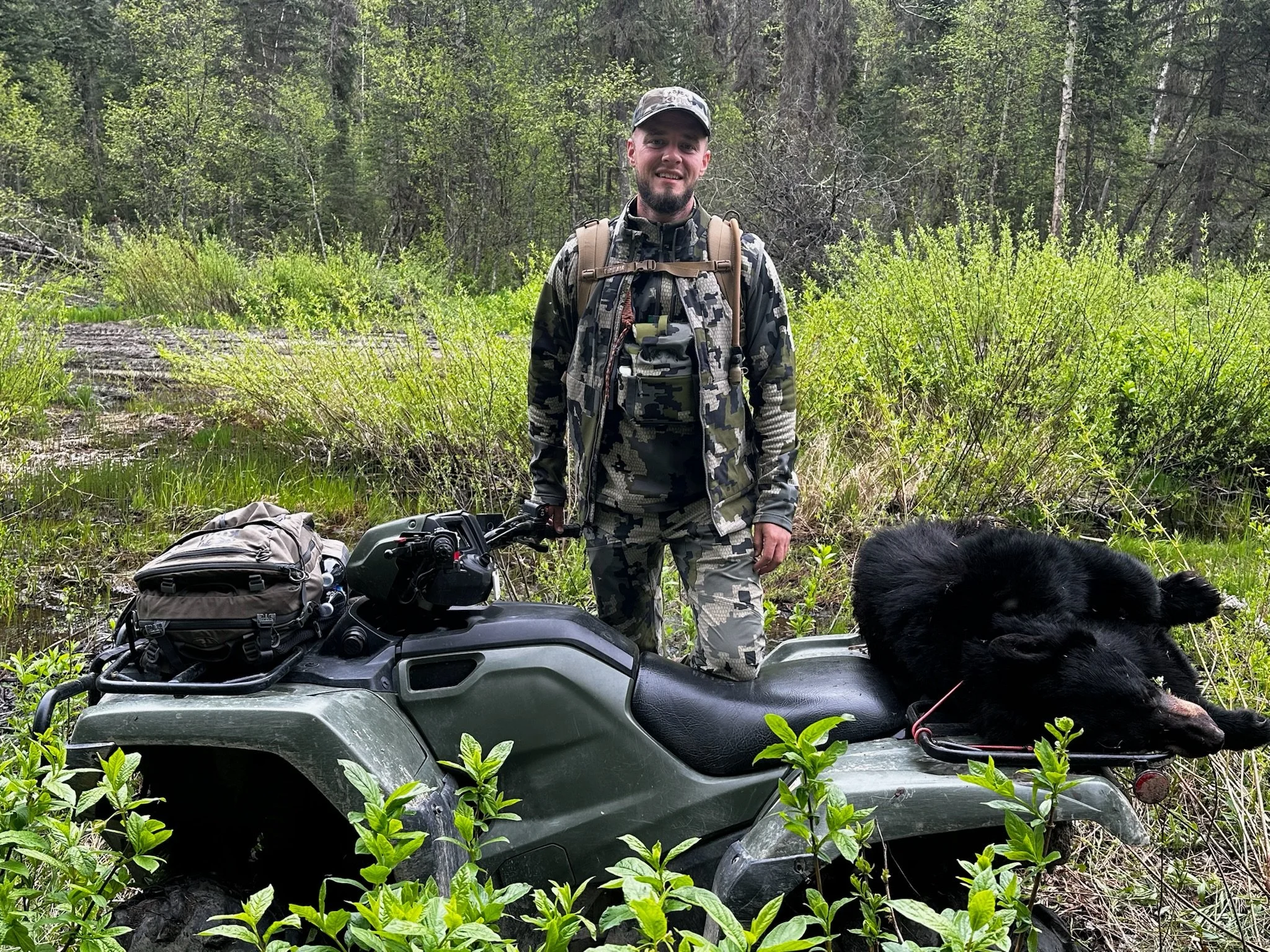 A man in camouflage clothing standing outdoors on a trail in a forested area, with a black dog lying on an ATV.
