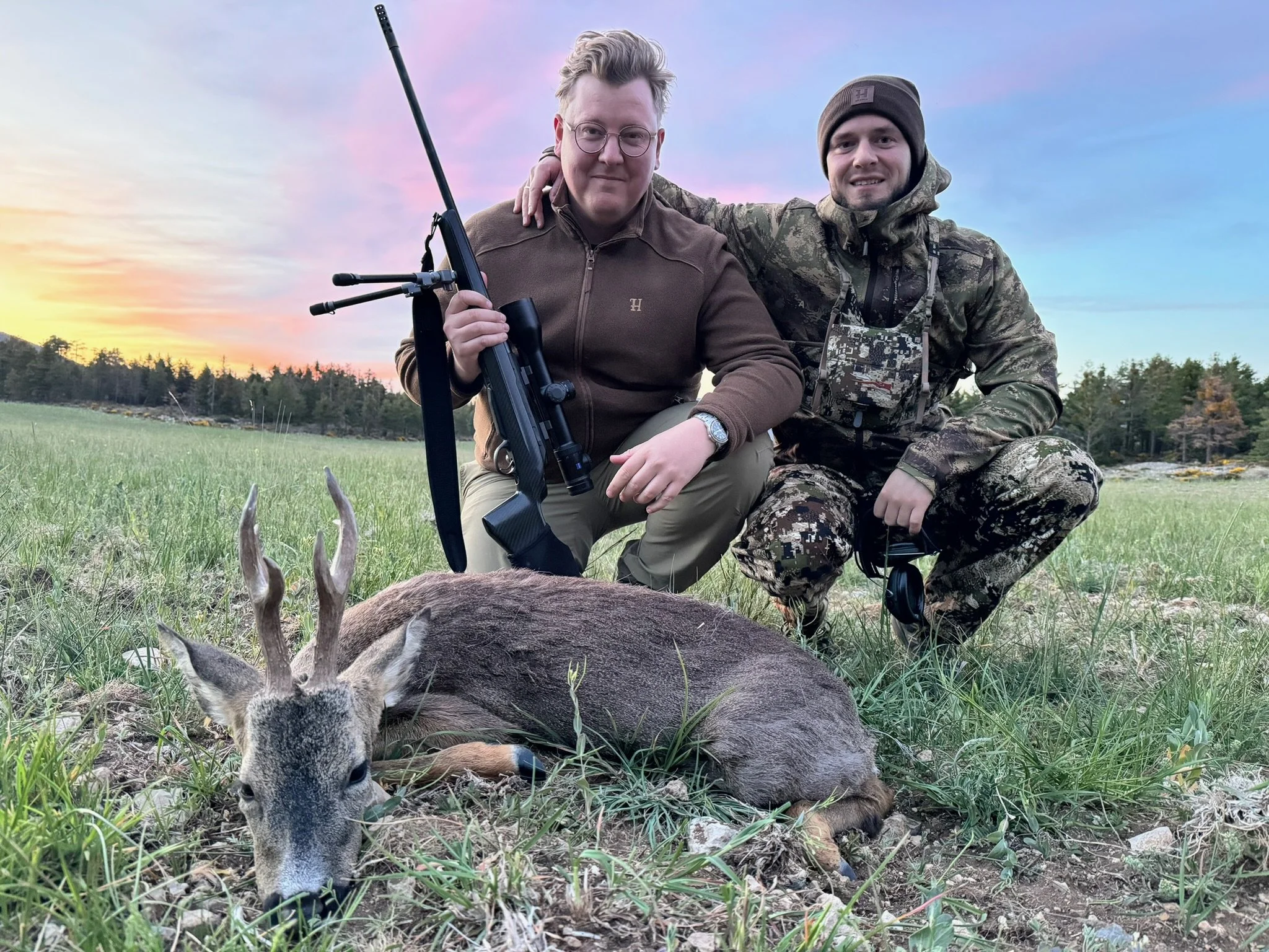 Two men in camouflage and outdoor clothing kneel beside a deer they appear to have hunted, with one holding a rifle. The deer is lying on the grass with broken antlers. They are outdoors during sunset, with a forest and a colorful sky in the backgrou