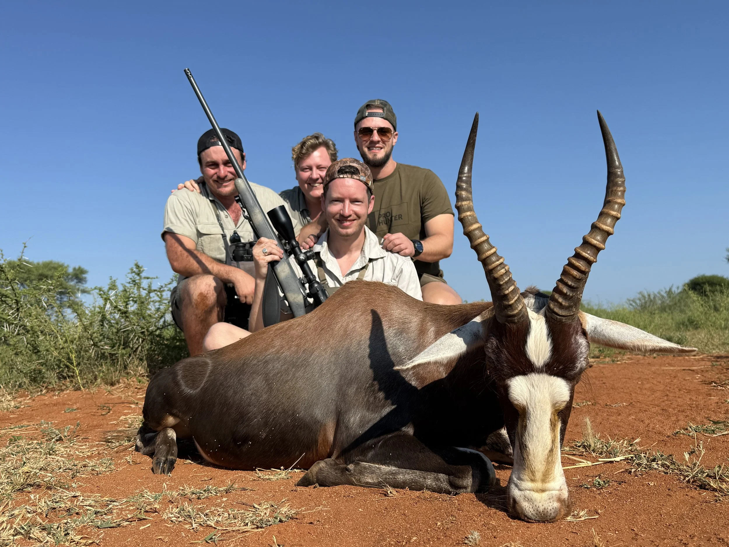 A group of four people smiling and posing behind an antelope with large, spiral horns that is lying on the ground in a desert landscape with blue sky and sparse vegetation.
