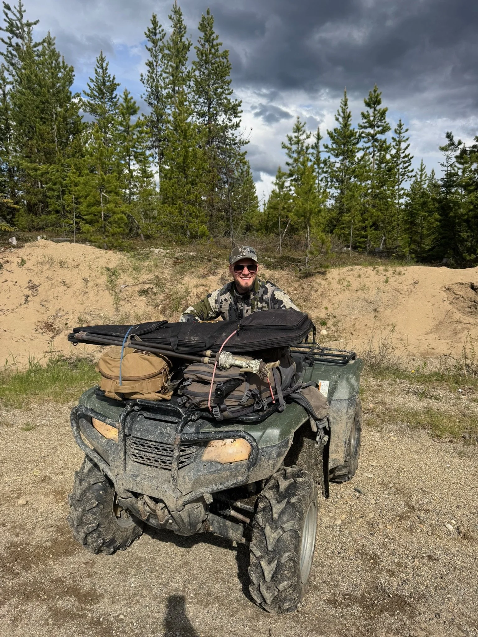 Smiling man in camouflage gear wearing sunglasses, sitting on a four-wheeler filled with gear and supplies, in a forested area under a partly cloudy sky.