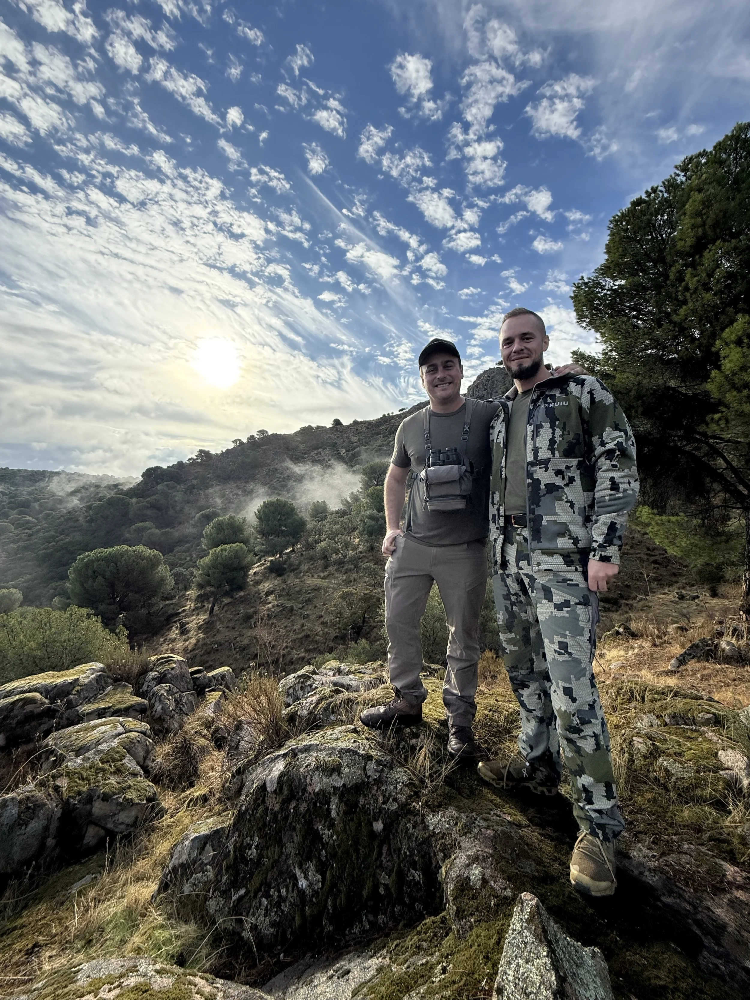 Two men stand on rocks in a mountainous landscape during sunset with a partly cloudy sky.