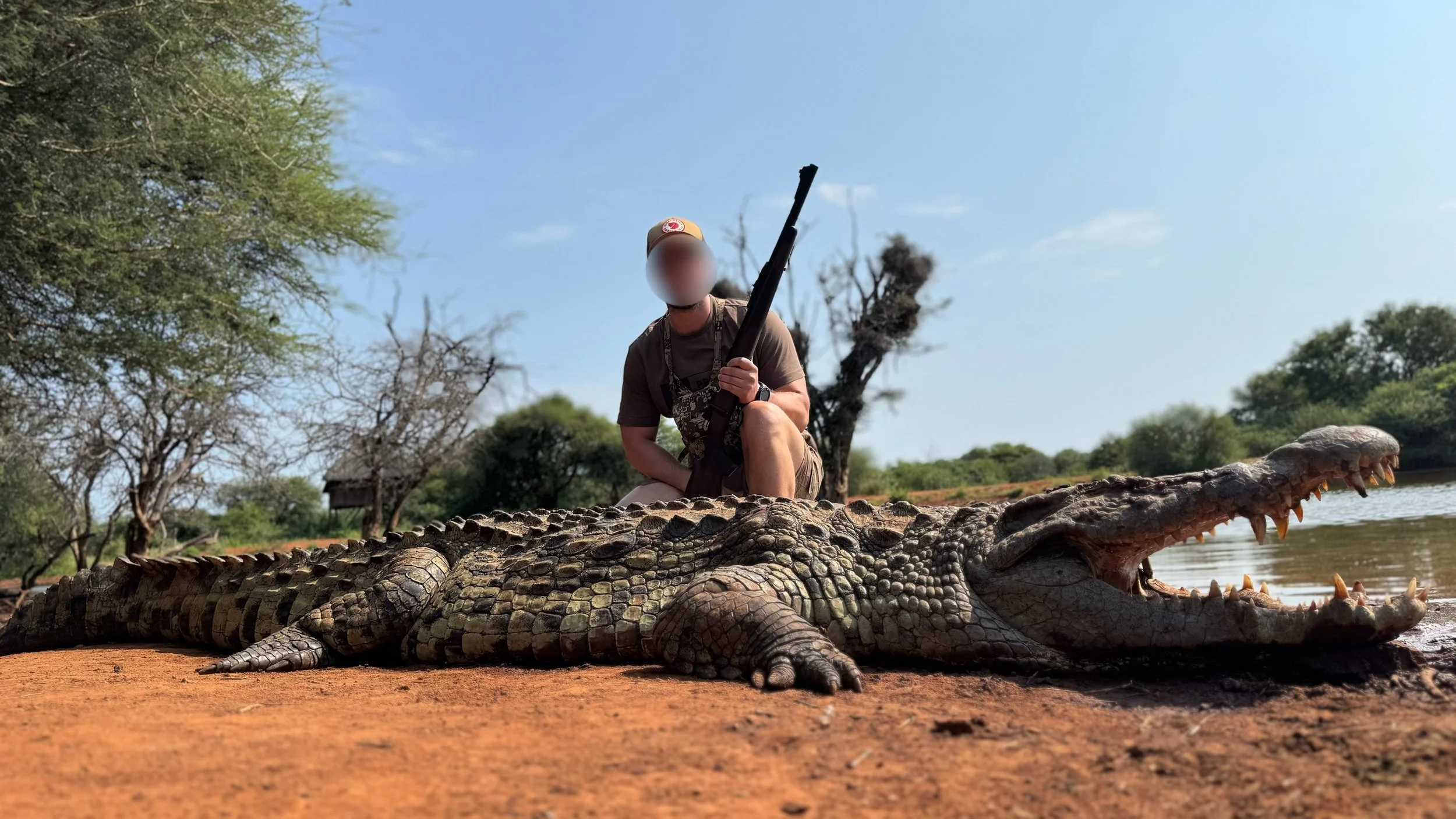 A person kneeling on the ground with a rifle next to a large crocodile lying on the ground near a body of water, surrounded by trees.