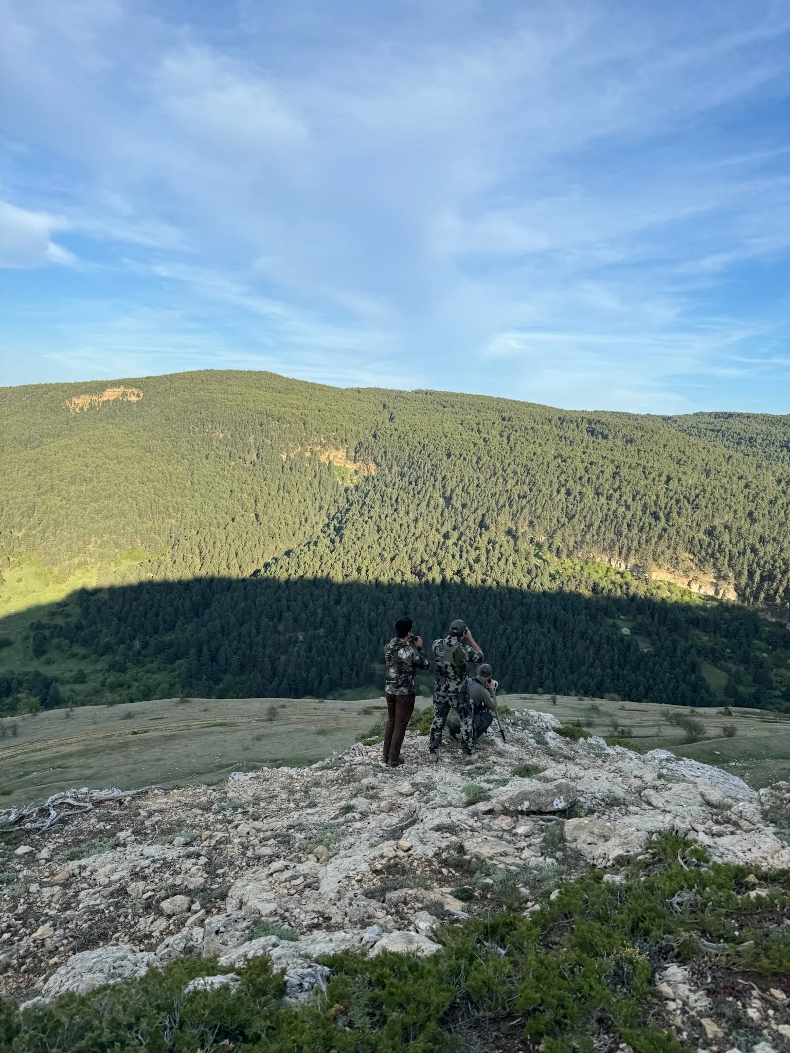 Four people in camouflage clothing standing on rocky terrain on a hill, observing the valley and forested mountains in the distance under a partly cloudy sky.
