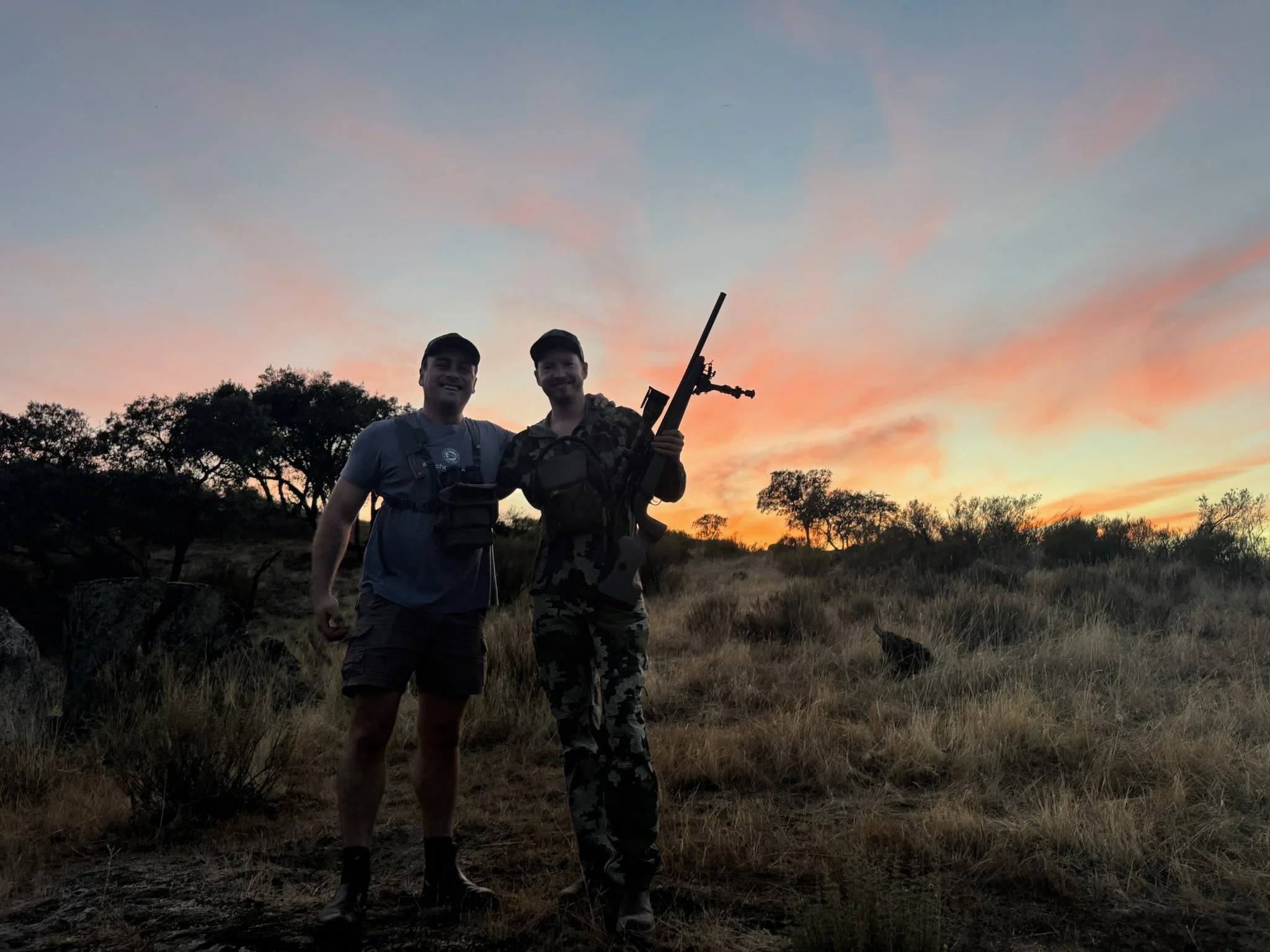 Two people, one in camouflage military gear and the other in casual outdoor clothing, stand together with arms around each other and smile at sunset in a grassy landscape with scattered trees and a dog in the background.